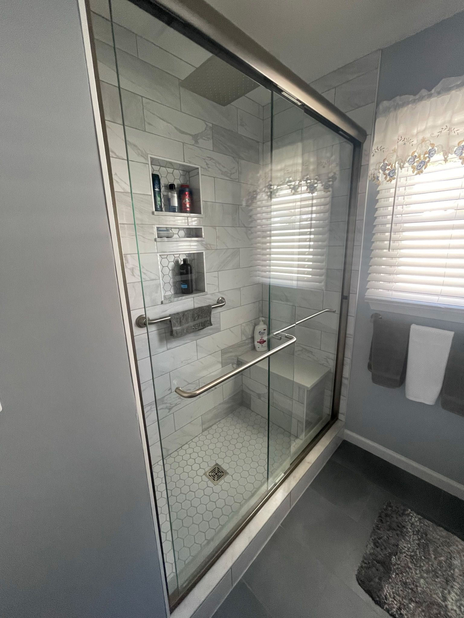 Modern bathroom shower with glass door, white tile, and wall-mounted shelves beside a window.