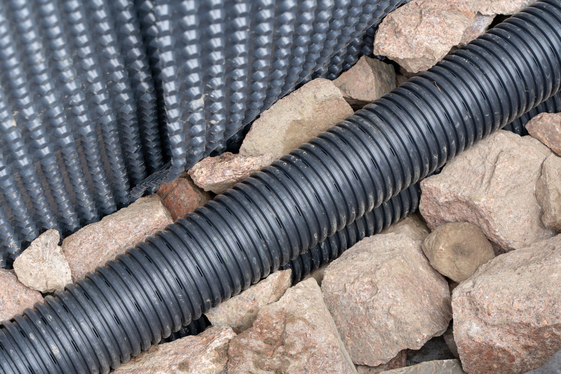 Tuyau de drainage noir ondulé entouré de pierres, à côté d'une barrière noire texturée.