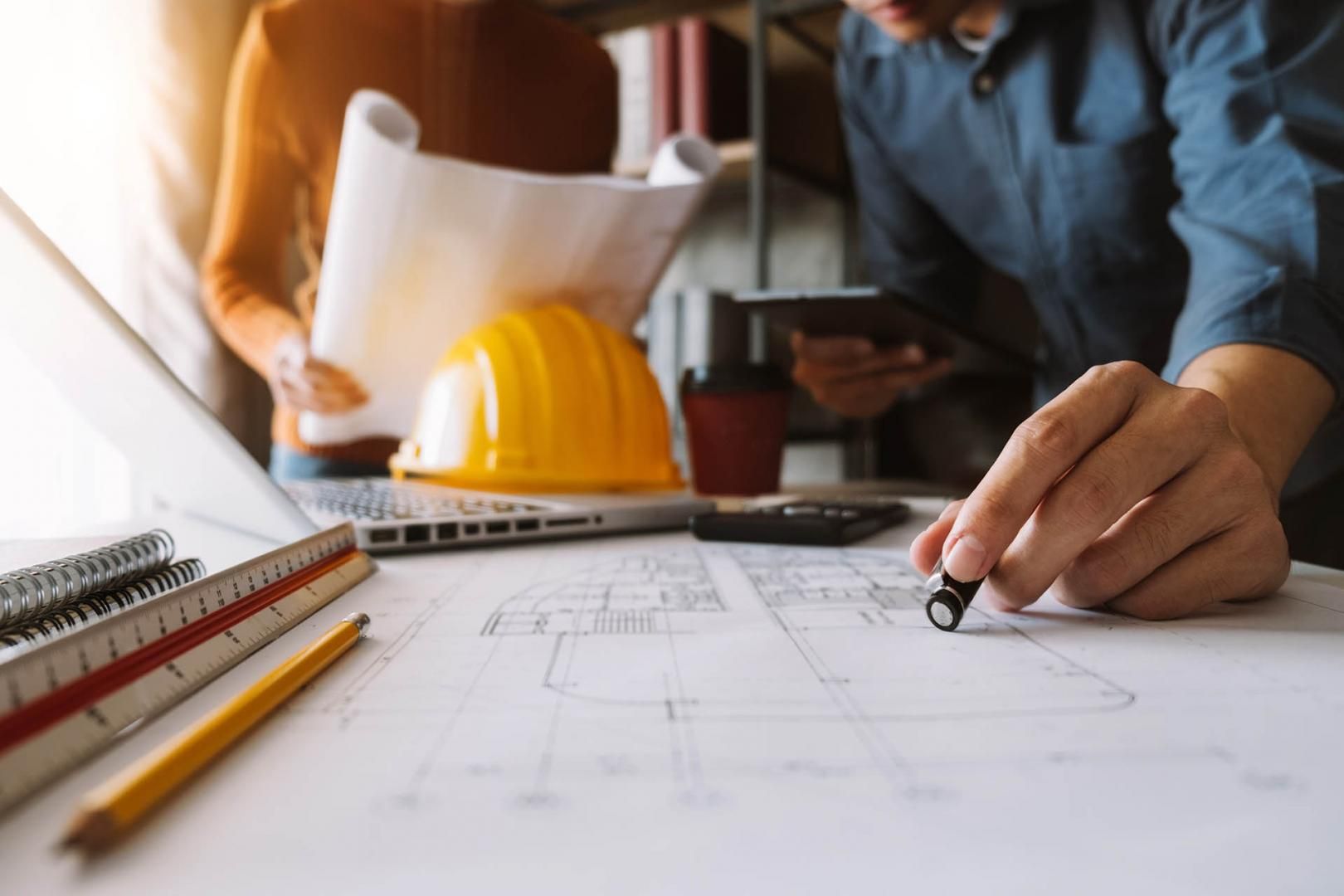 Deux personnes examinent des plans, projet de construction, avec un ordinateur portable, un casque de chantier et une règle.
