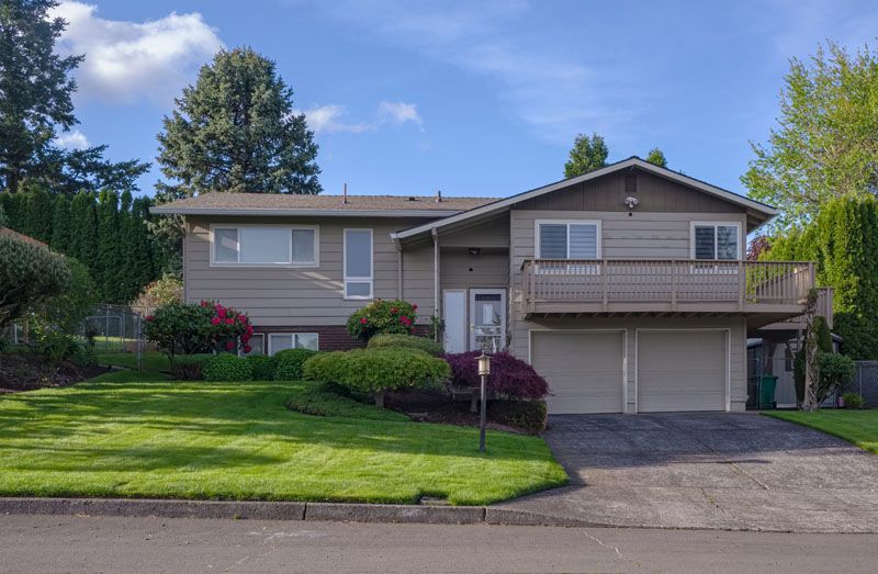 Maison à deux étages avec un revêtement extérieur beige, un balcon et un garage double, sur une pelouse verte sous un ciel bleu.
