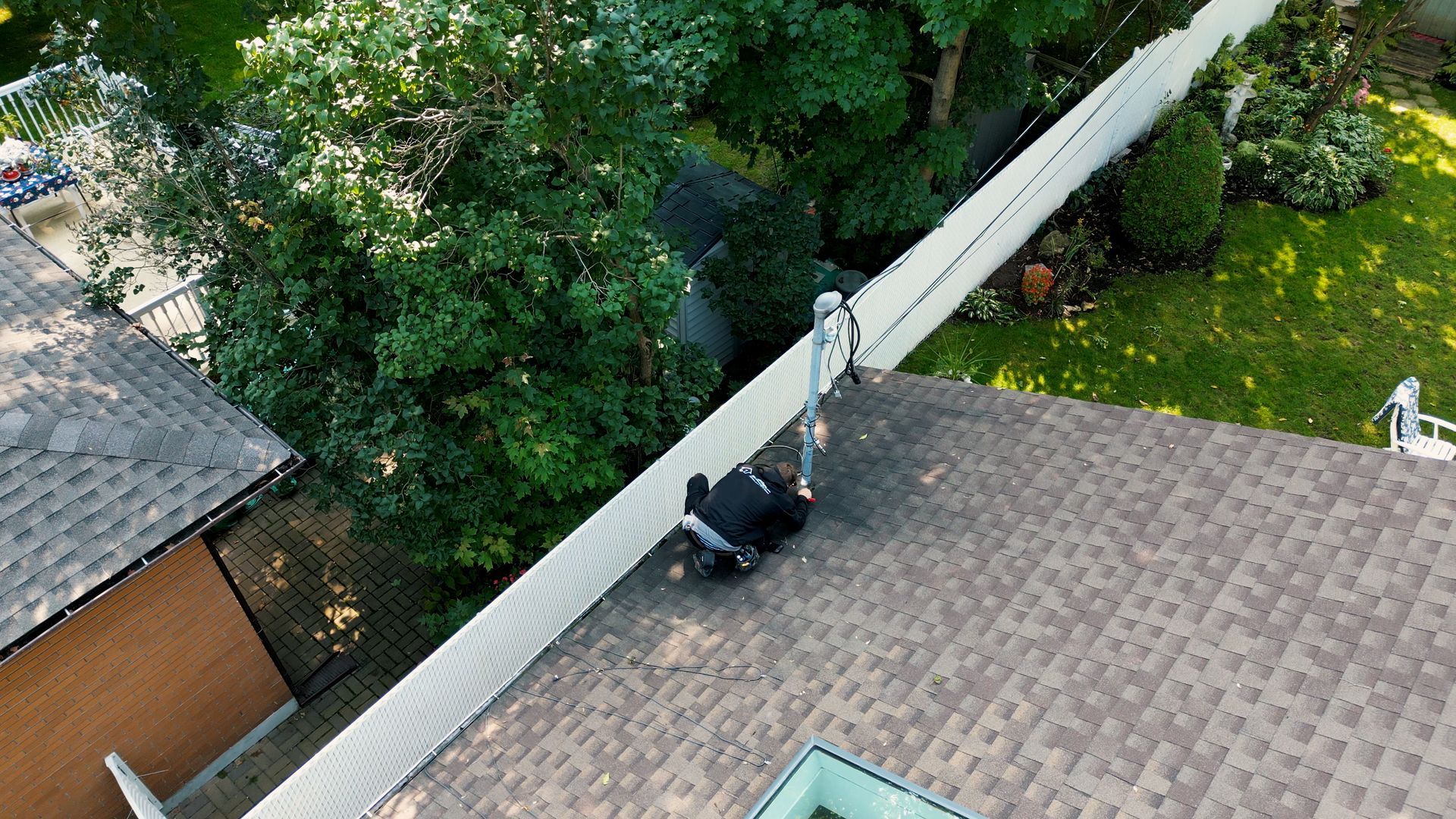 Un technicien vêtu de sombre est agenouillé sur un toit de bardeaux, travaillant sur un mât de service public installé près du bord.
