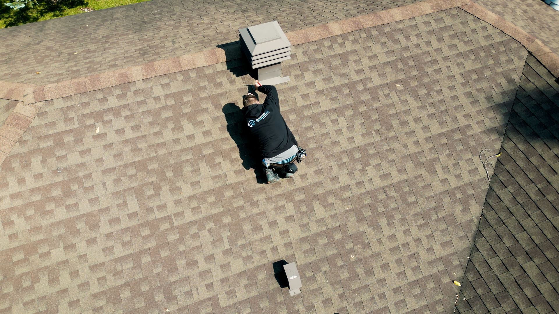 Une personne vêtue d'une veste sombre est agenouillée sur le toit d'une maison, travaillant près d'une grille d'aération.