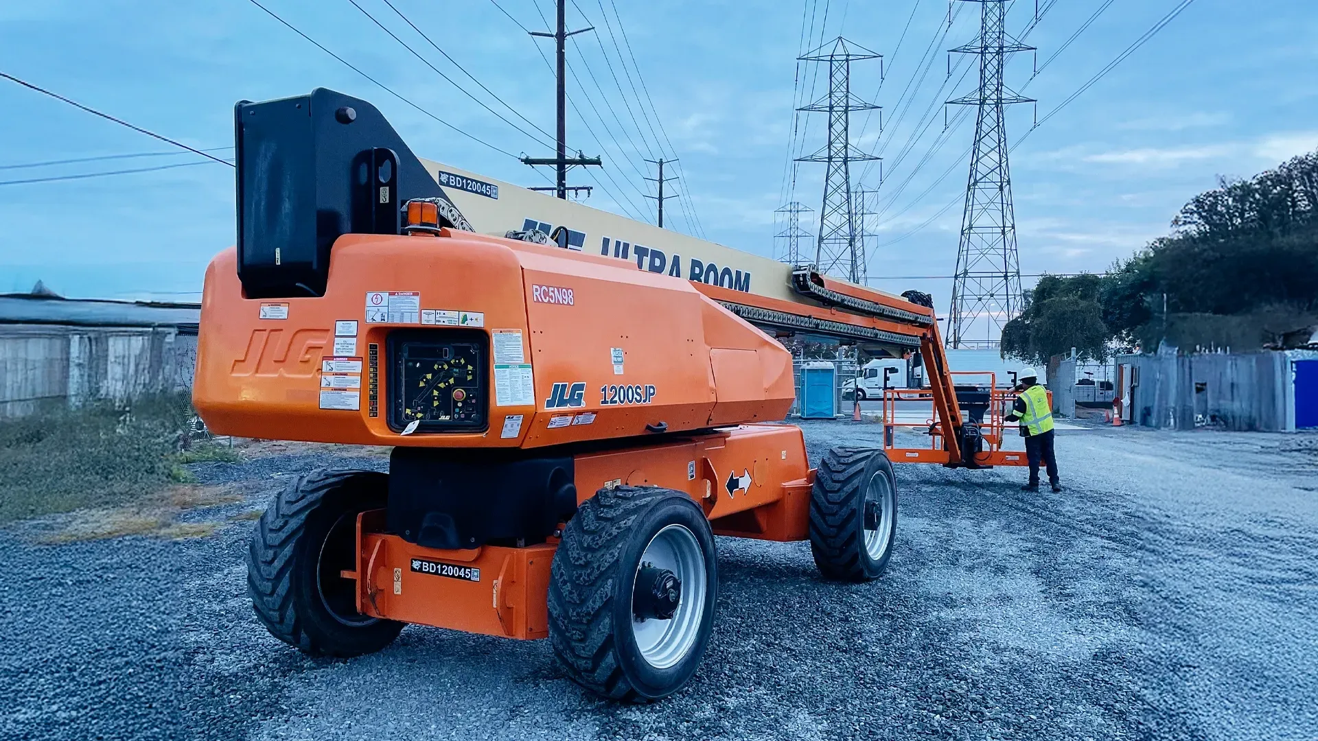 A large orange boom lift staged at a utility work site with power lines in the background and a crew