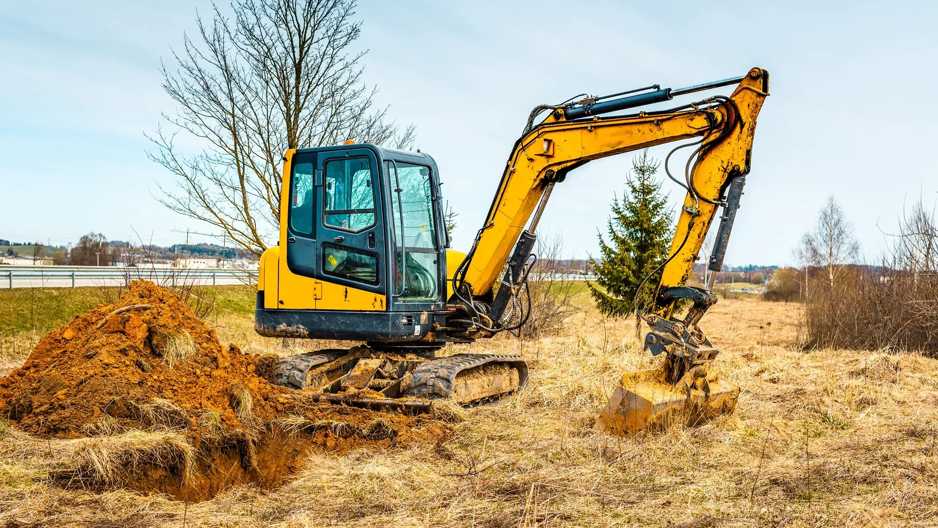 A compact yellow excavator parked on a grassy field beside a fresh dirt pile under a cloudy sky.