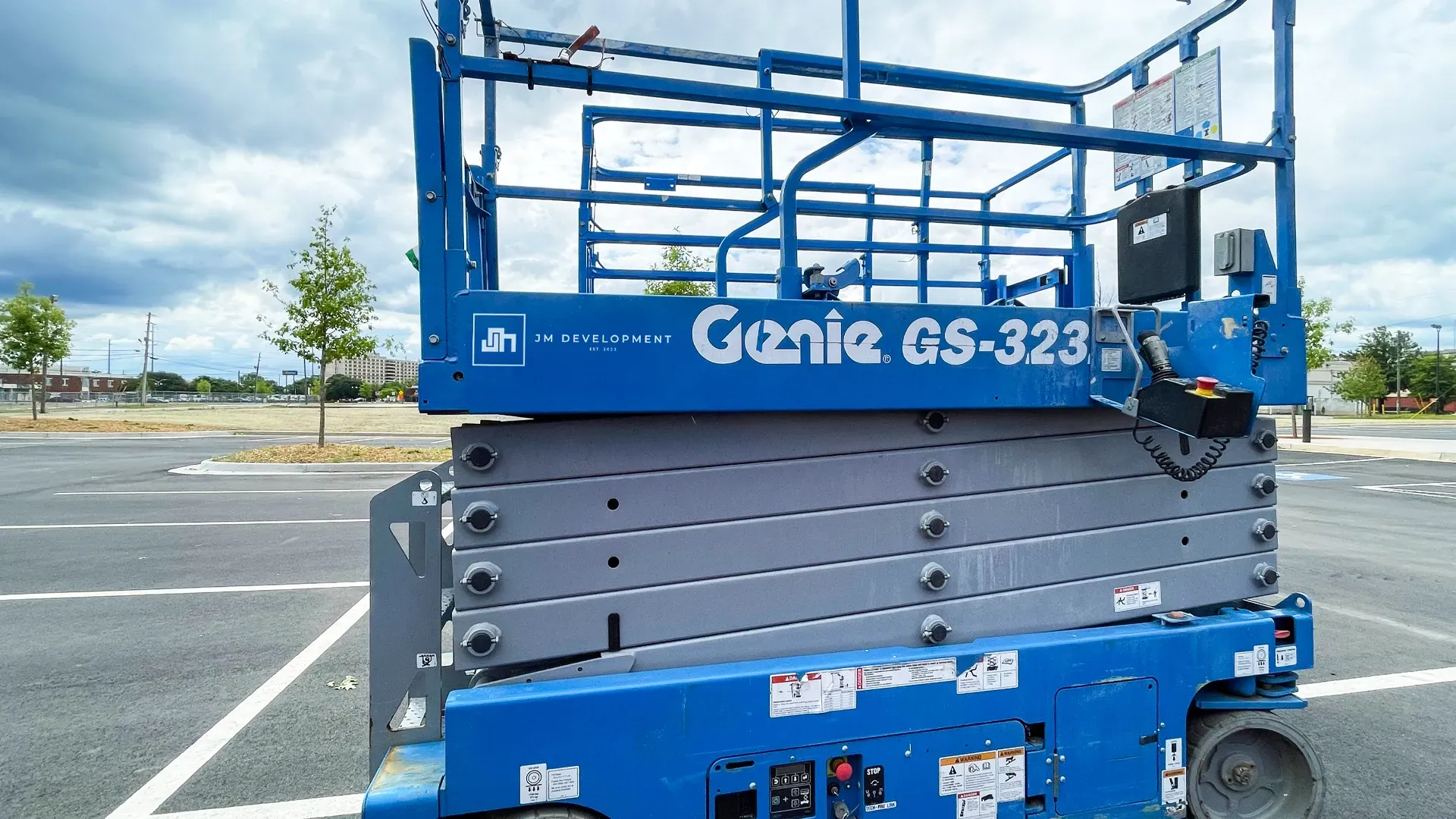 A parked scissor lift with JM Development branding, in a large open lot under cloudy skies.
