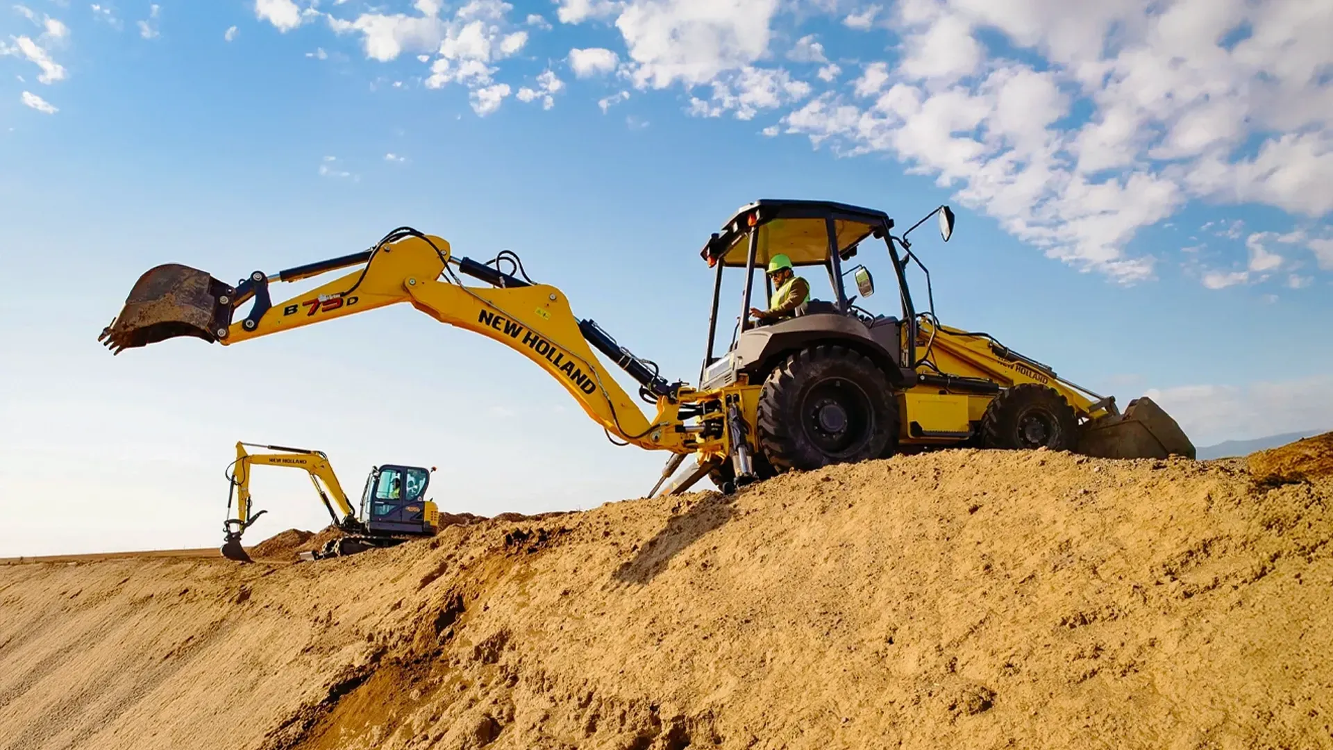 A yellow New Holland backhoe loader operating on a dirt slope under a bright blue sky, symbolizing J