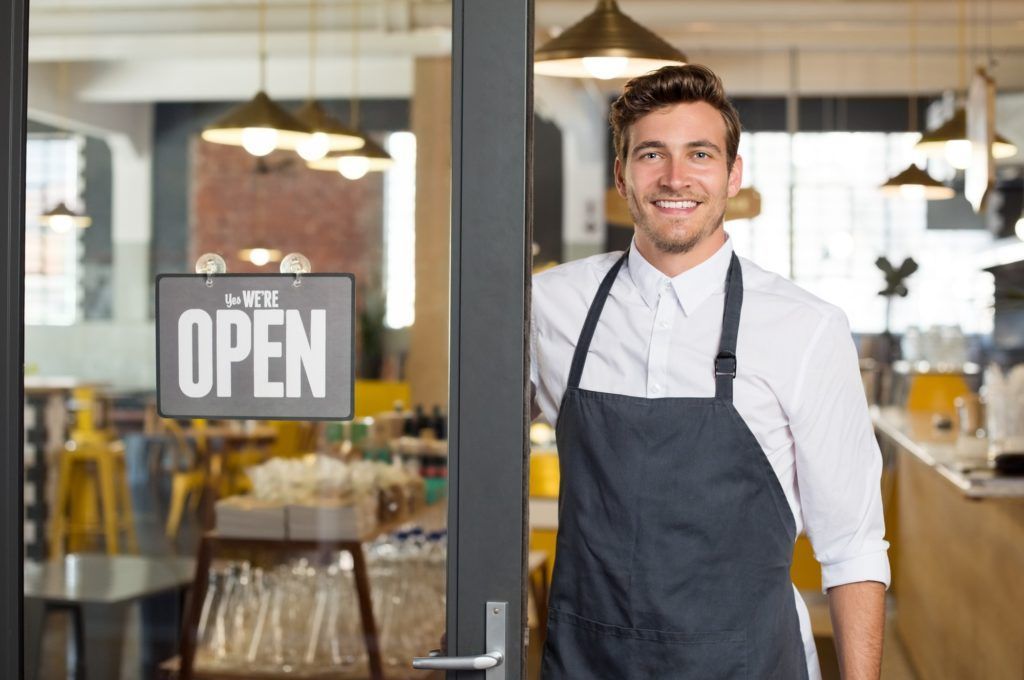 A man in an apron is standing in front of an open restaurant door.