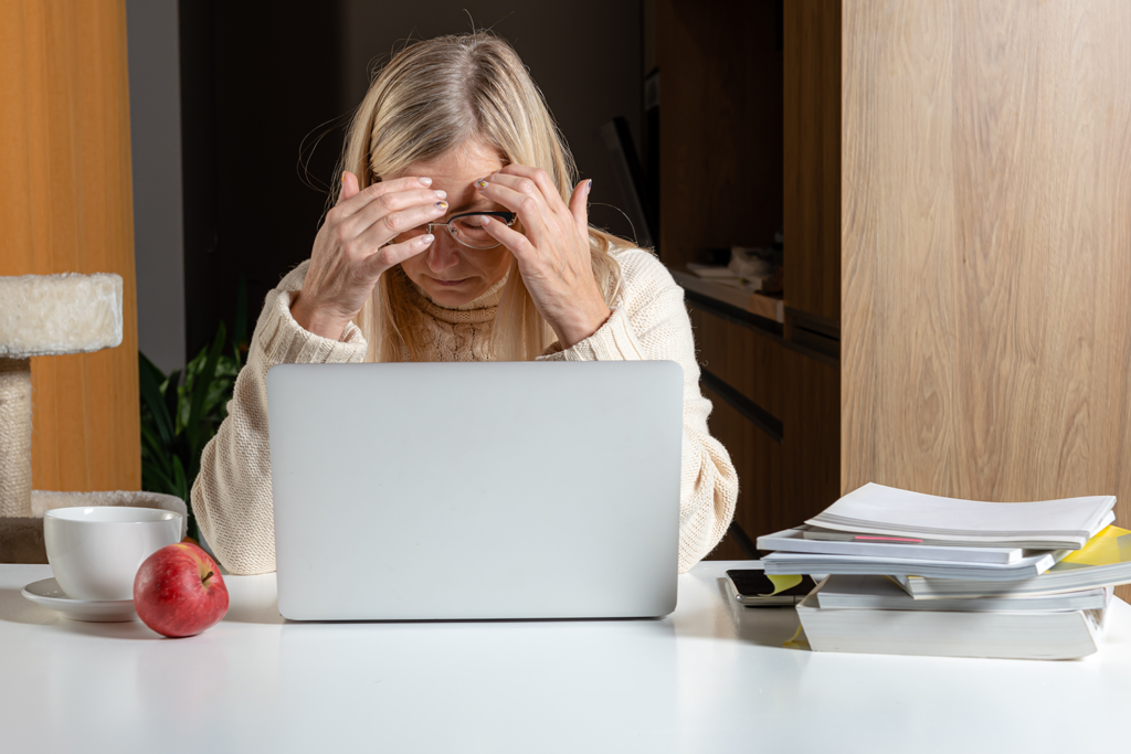 A woman is sitting at a desk with a laptop computer and rubbing her eyes.