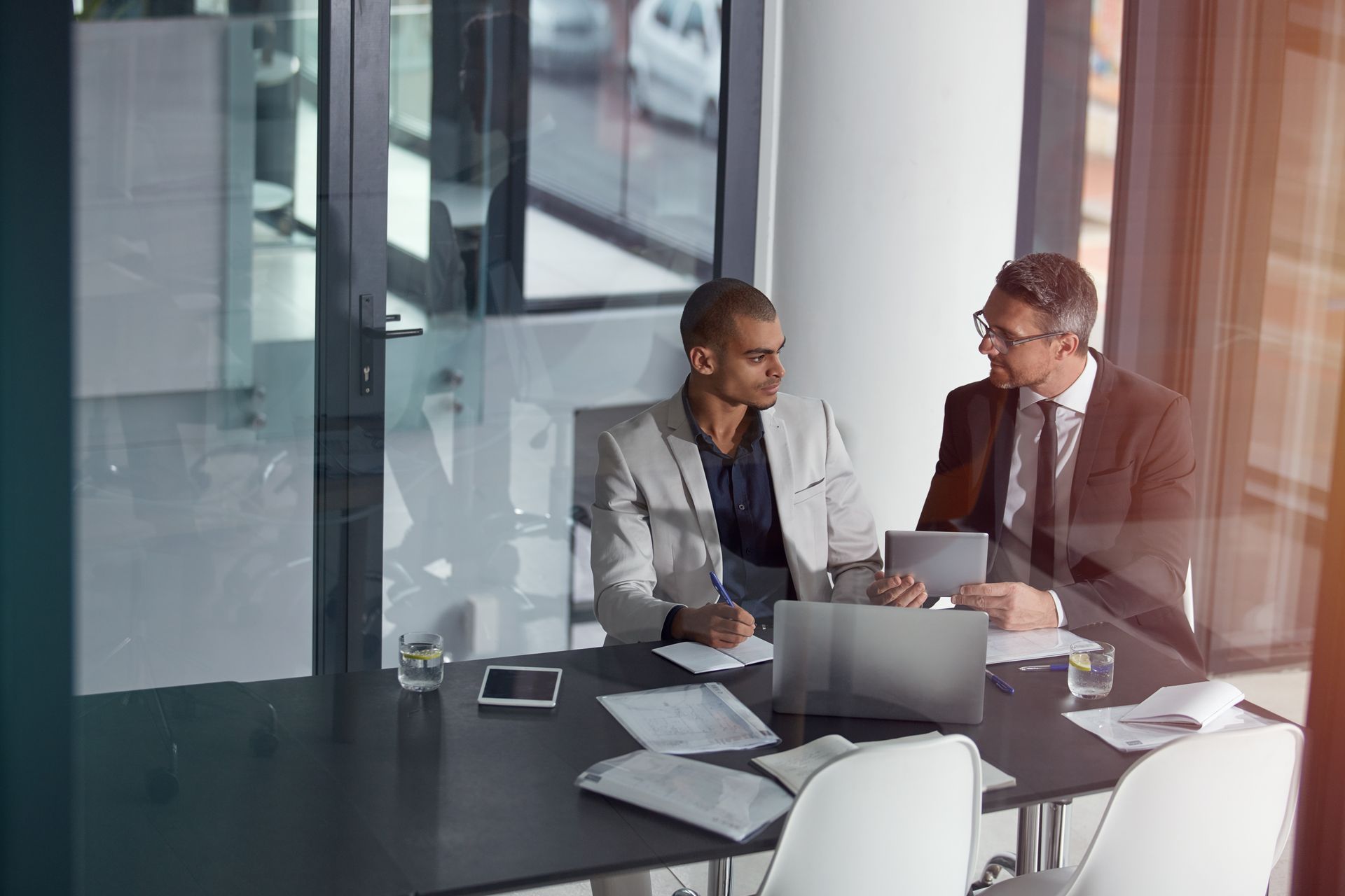 Two men are sitting at a table in a conference room looking at a laptop.