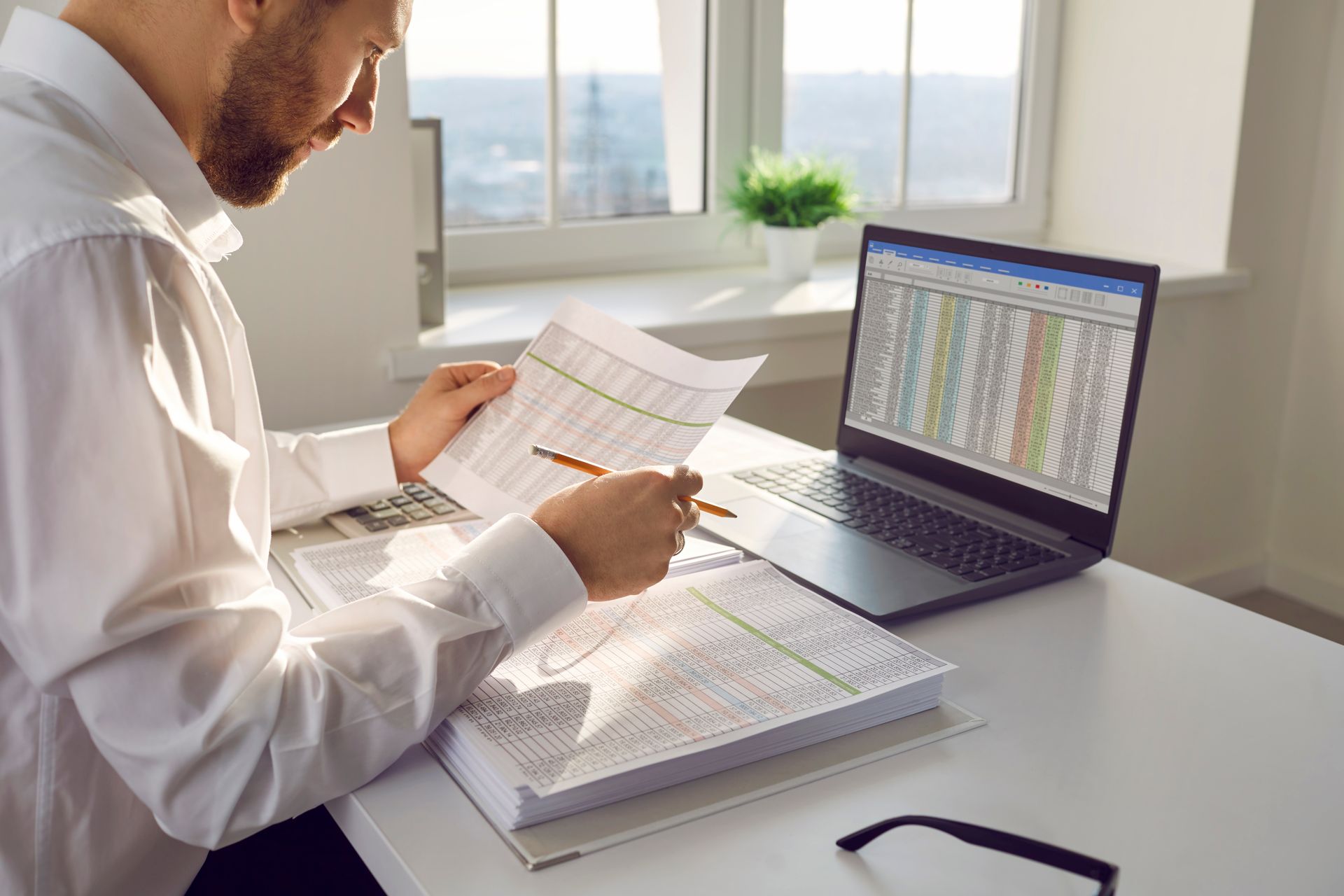 A man is sitting at a desk with a laptop and a stack of papers.
