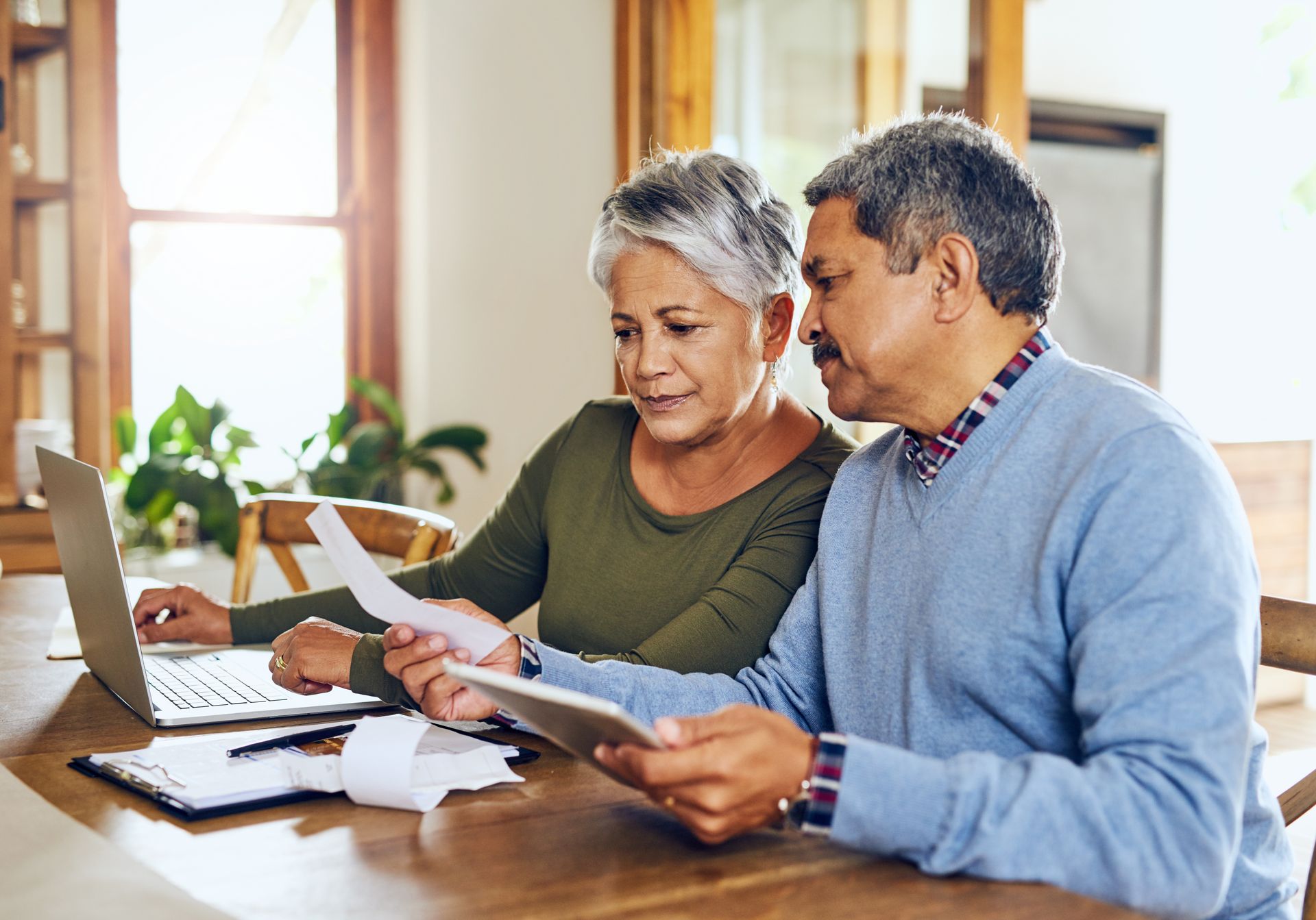 An elderly couple is sitting at a table looking at papers and a laptop.