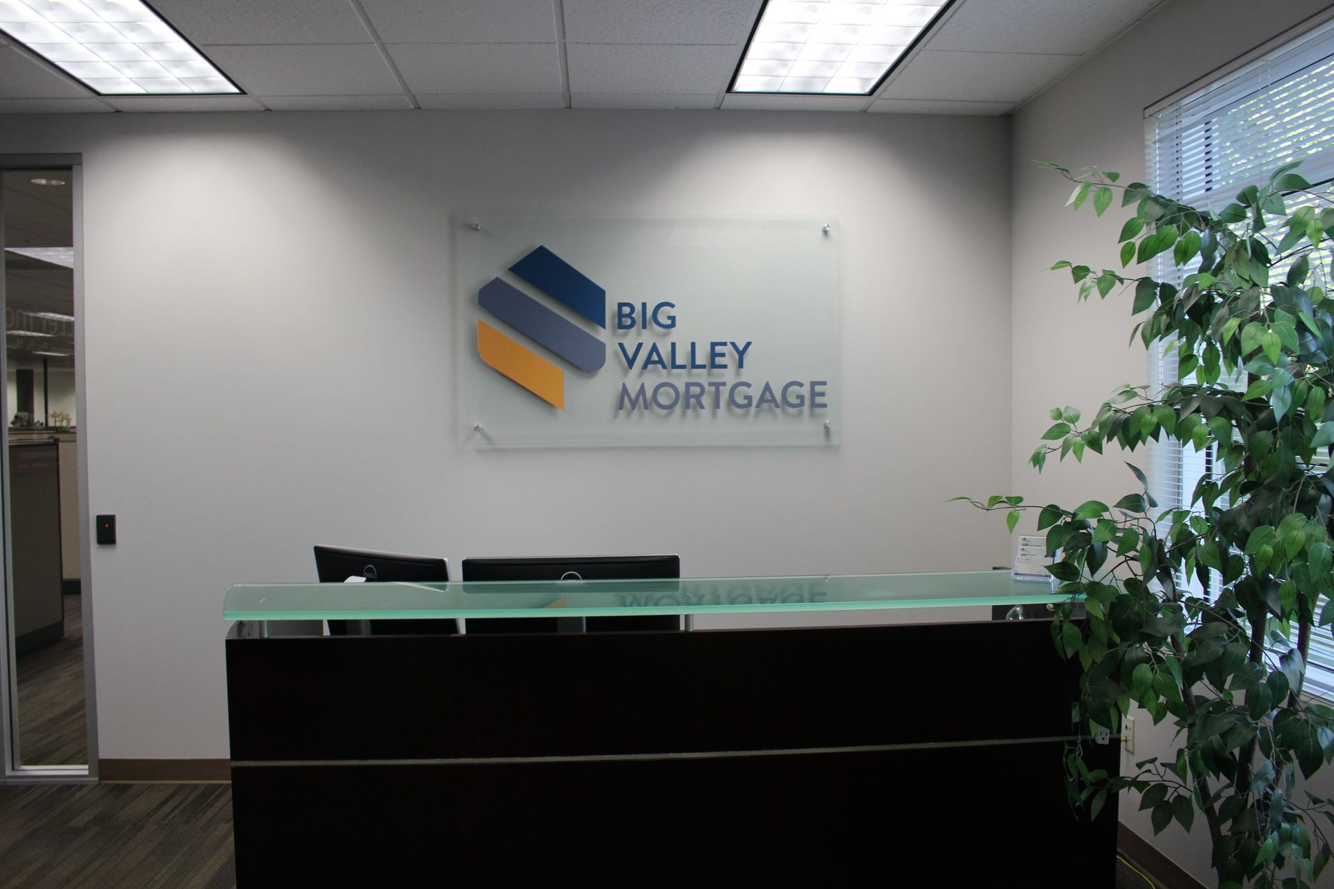 A modern office reception area with a glass desk and the Big Valley Mortgage logo mounted on the wall behind it.