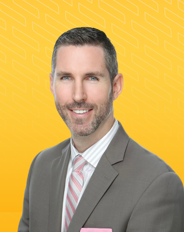 Headshot of Phil Rasmussen in a gray suit with a pink tie and pocket square, smiling against a bright yellow background.