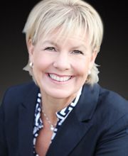 Headshot of Kristy Ruhkala wearing a dark blazer and patterned blouse, smiling against a dark gradient background.