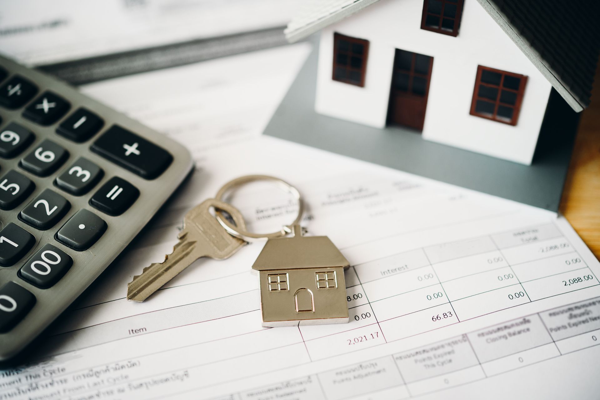 Calculator, house key, miniature house, and paperwork on a desk; financial planning concept.