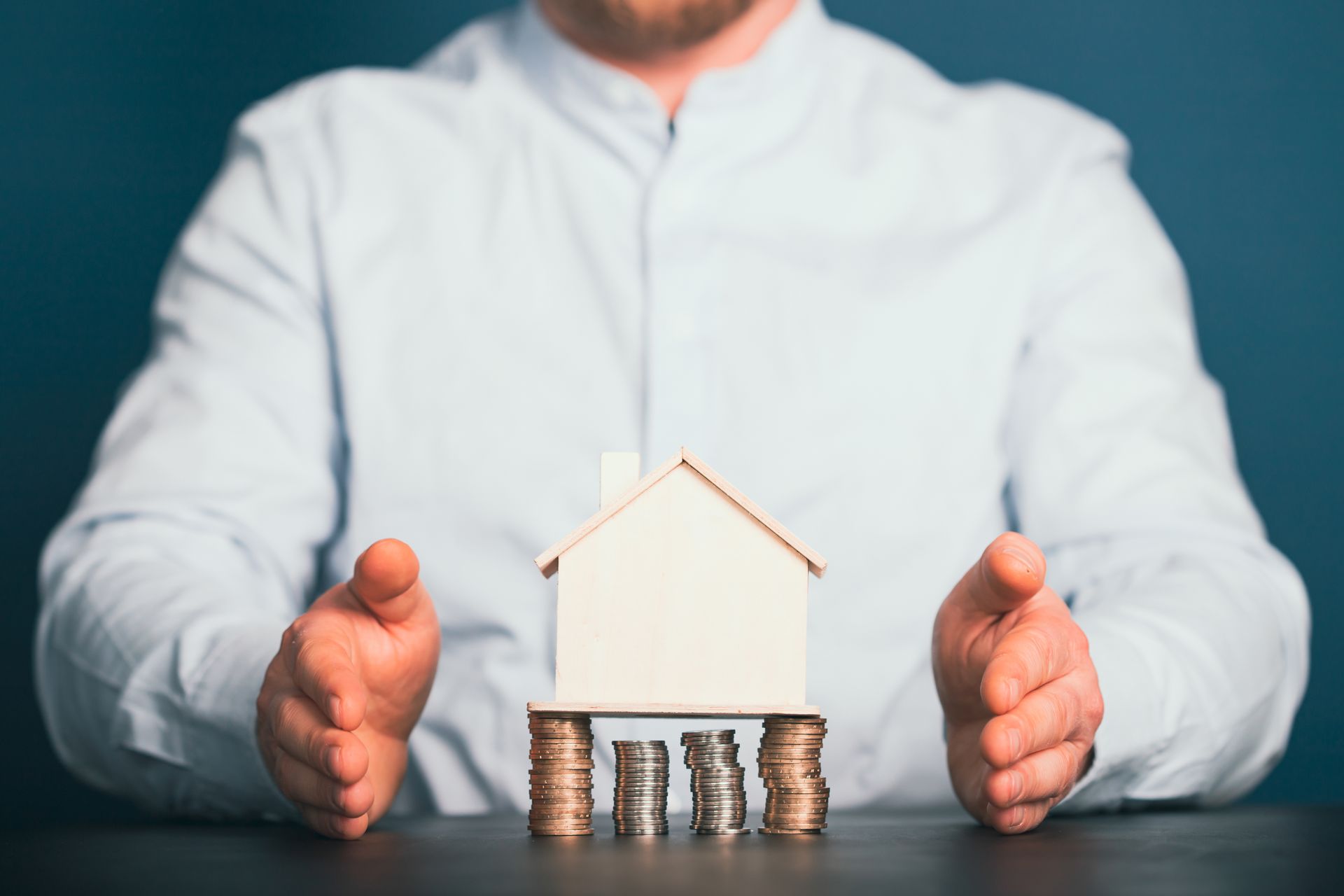 Person's hands protecting a model house perched on stacks of coins, against a blue background.