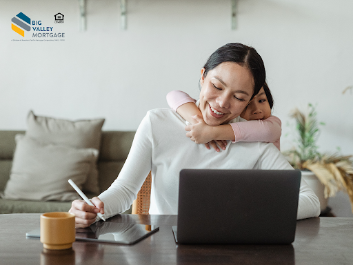 Woman working on laptop with daughter hugging her. Indoors. Smiling.
