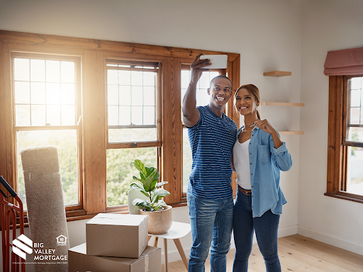 Couple takes selfie in a new home, smiling and holding a key. Boxes and plant sit on the floor.