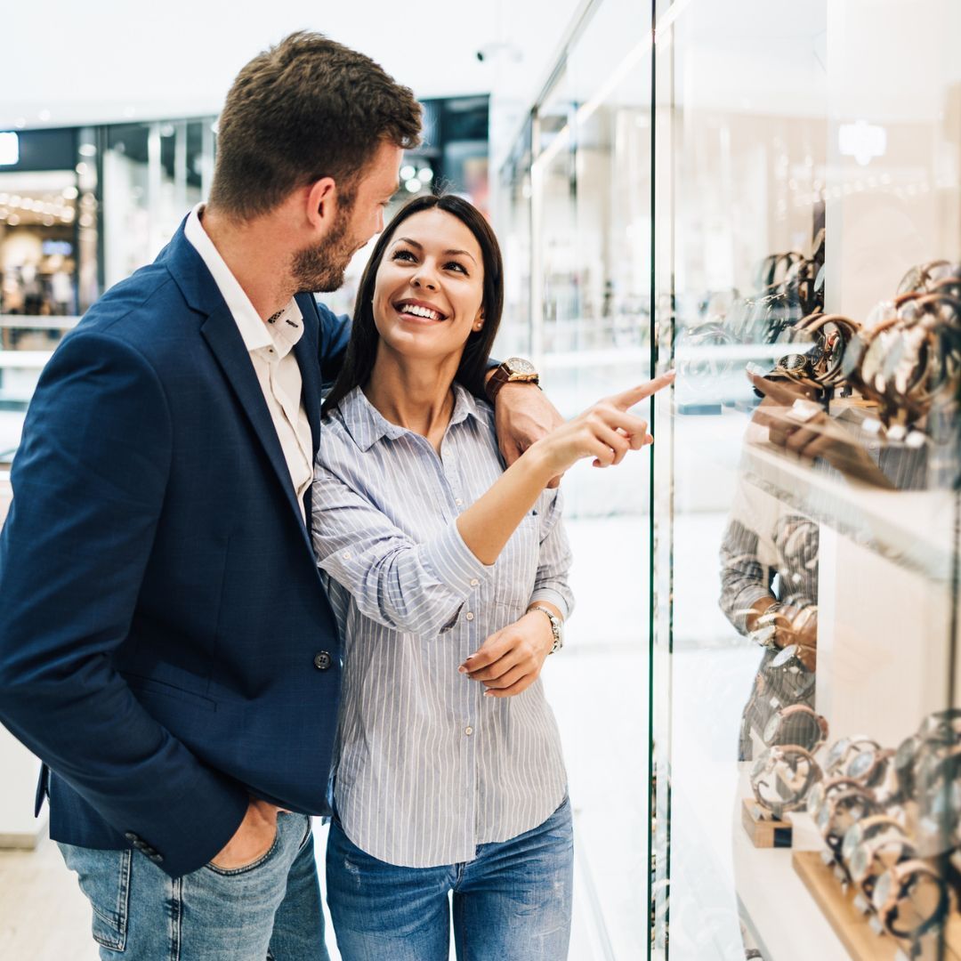 Couple looking at watches in a store. Woman points at a display while smiling. Man has arm around her.
