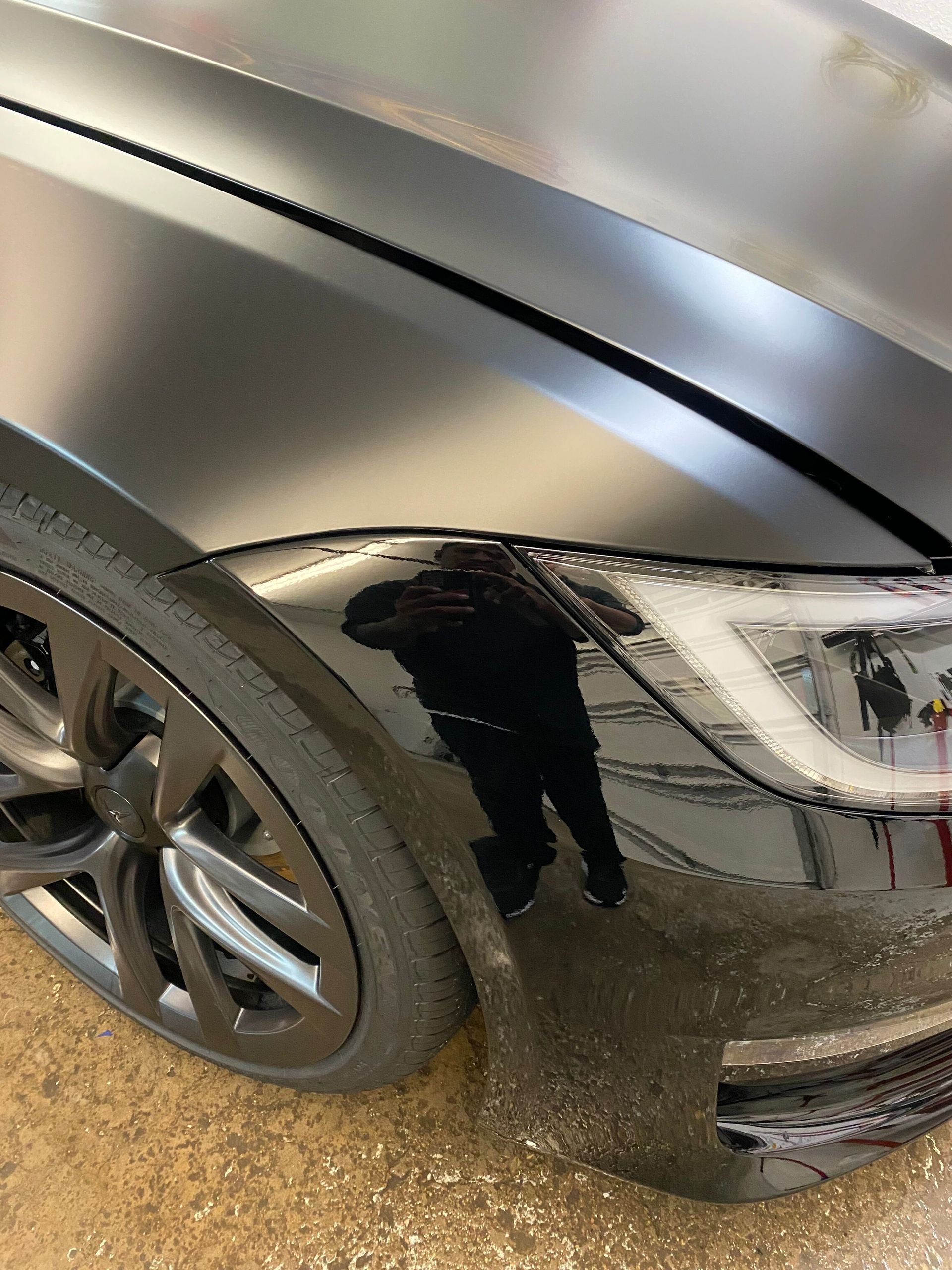 A black car is parked in a garage with a reflection of a person in the headlight.
