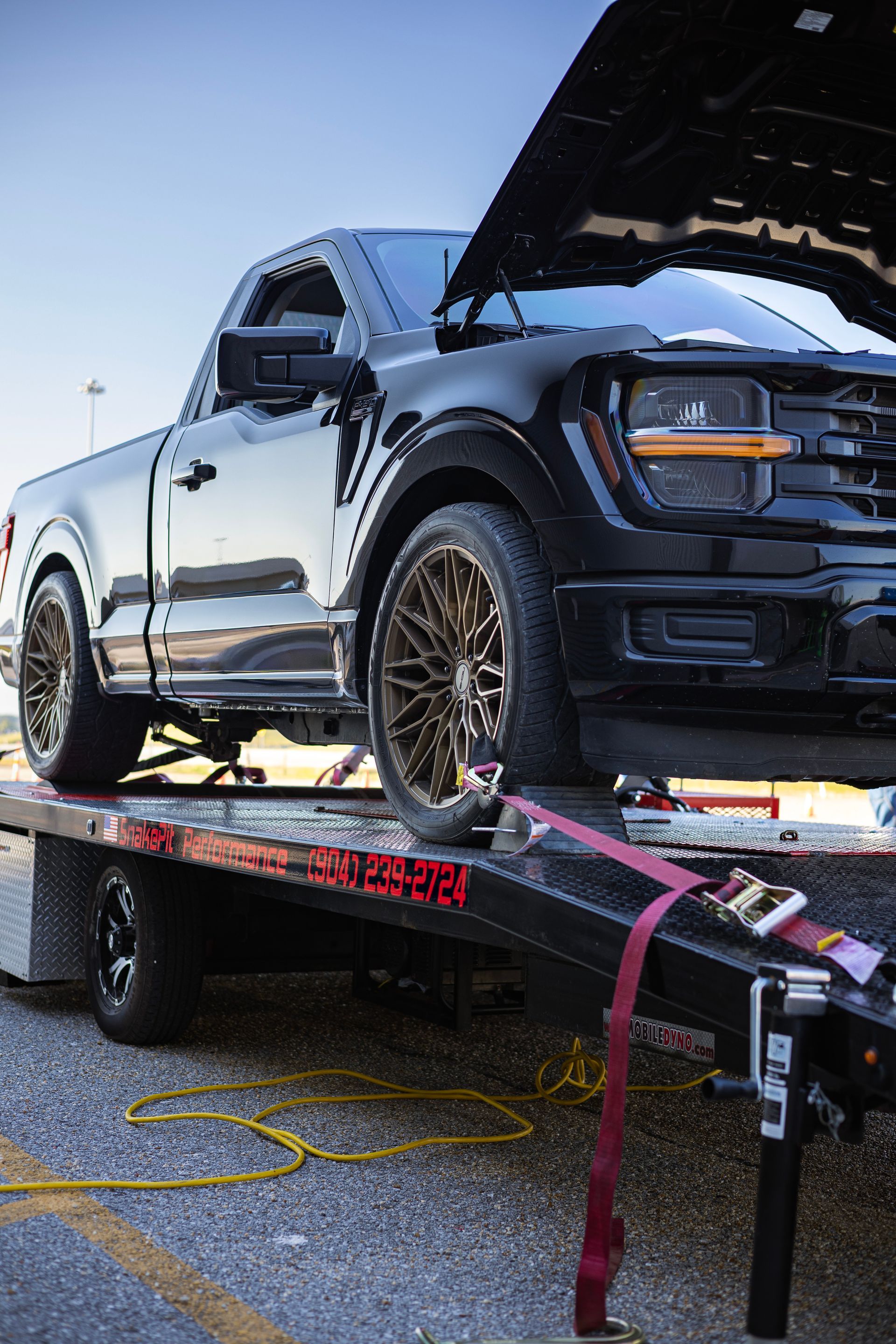 Black pickup truck on a trailer, hood open. Brown wheels, red strap, parked on asphalt.