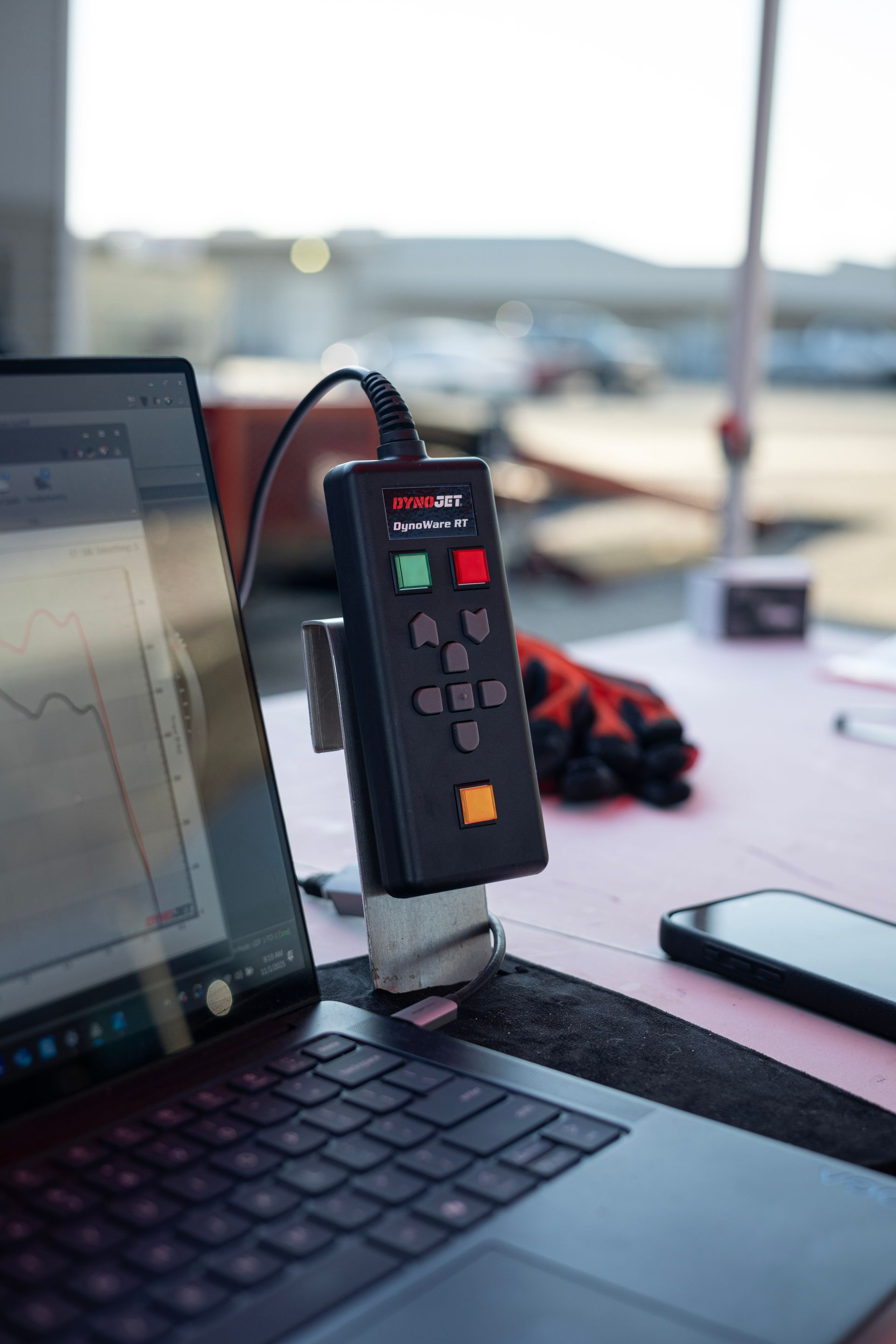 Laptop connected to a black control panel with buttons, mounted on a table. Gloves and phone are nearby.