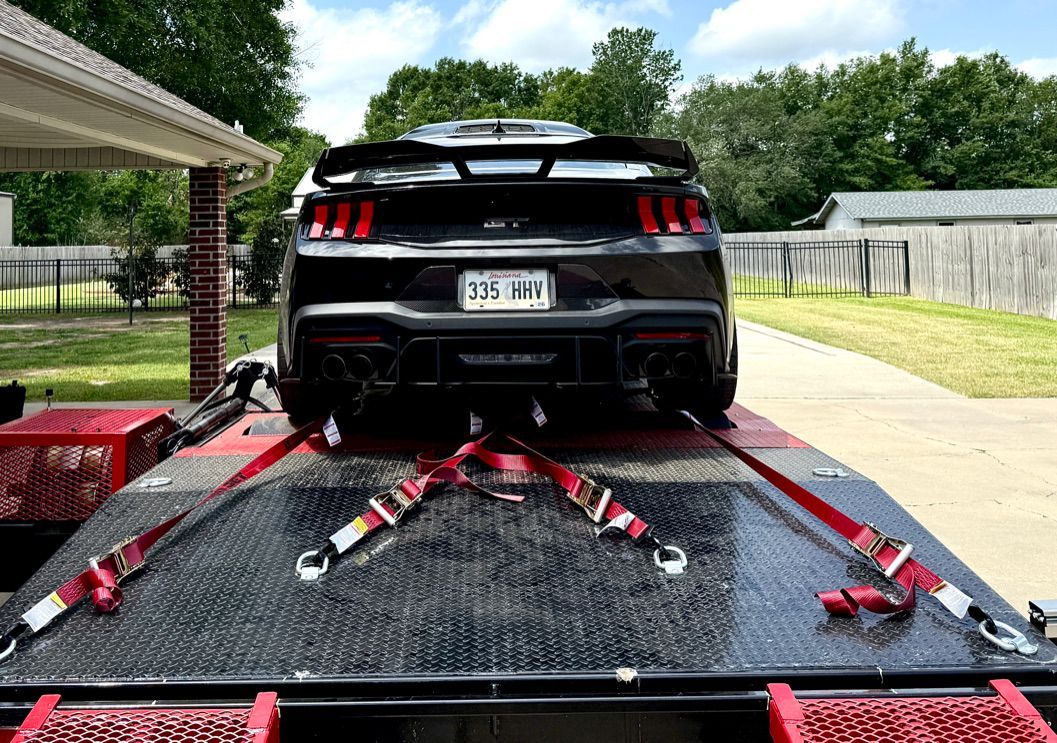 Black Ford Mustang secured with red straps on a black trailer, outdoors.