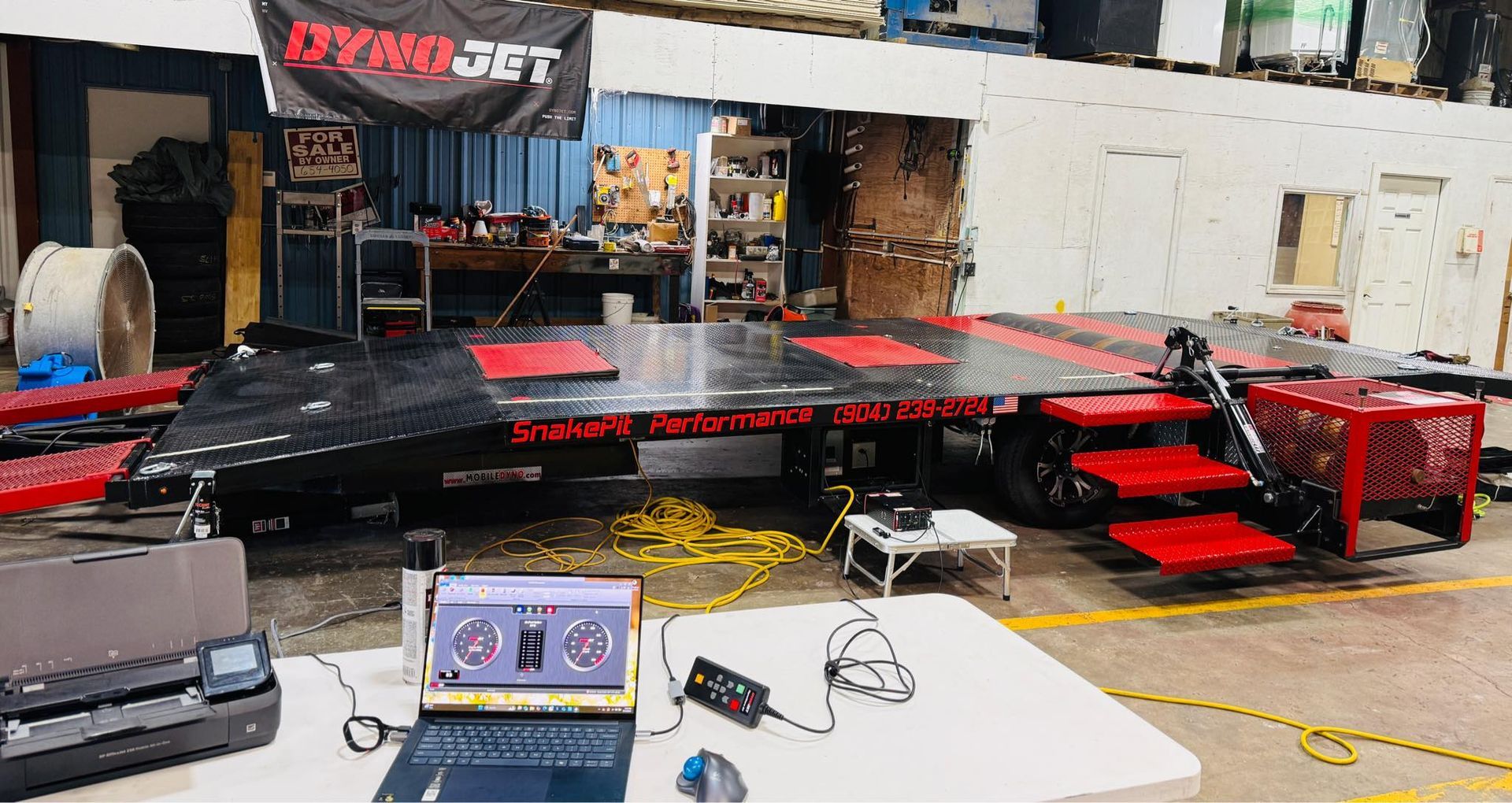 A dyno machine set up in a shop, with a laptop and equipment on a table, ready for engine performance testing.