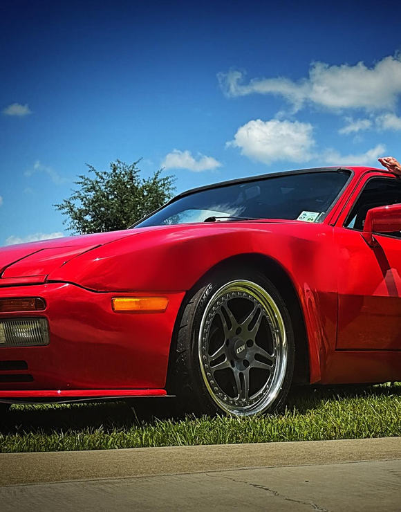 Red sports car parked on grass, blue sky with clouds in background.