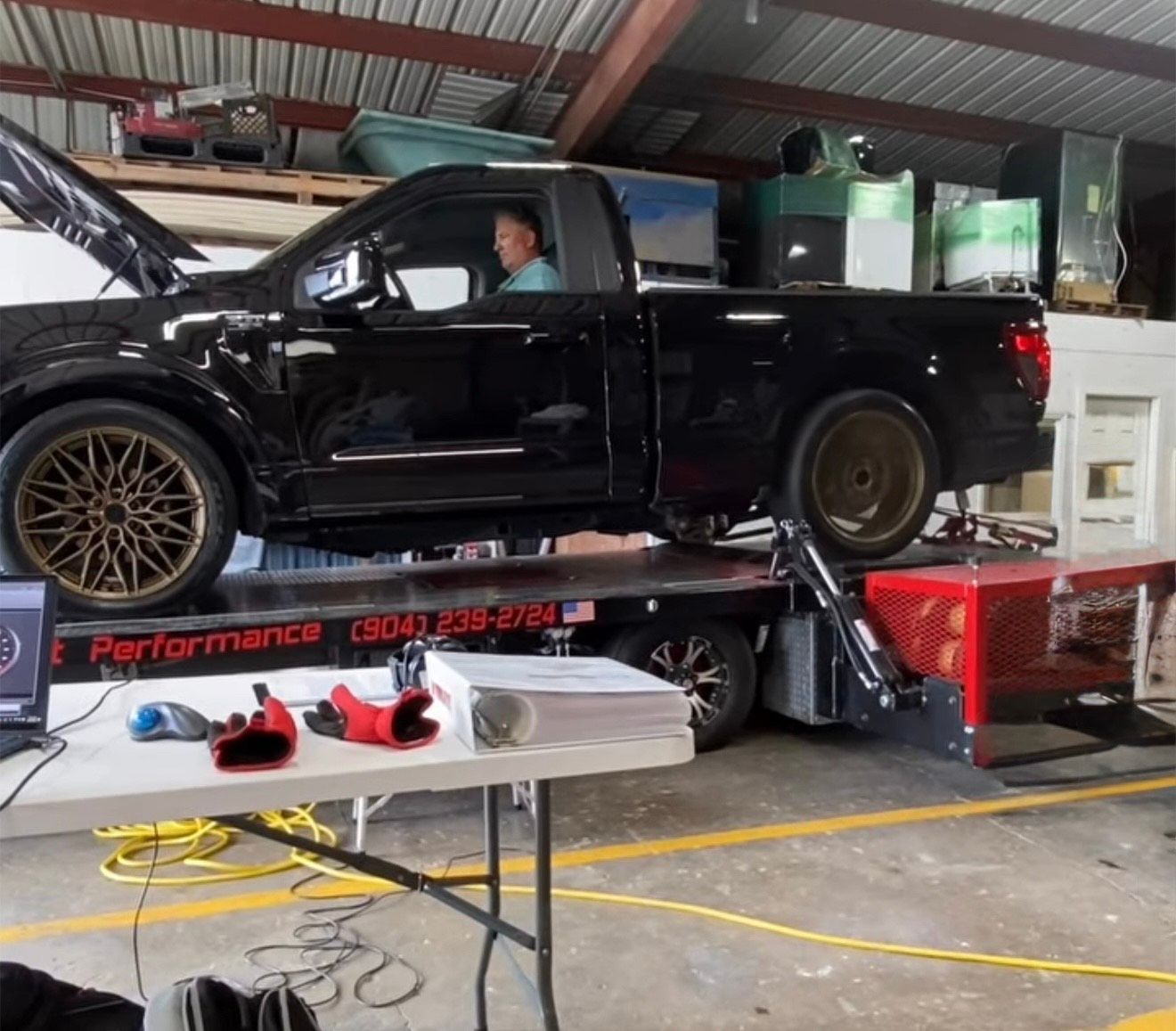 Black pickup truck on a dynamometer, hood open. Man inside. Shop setting with equipment.