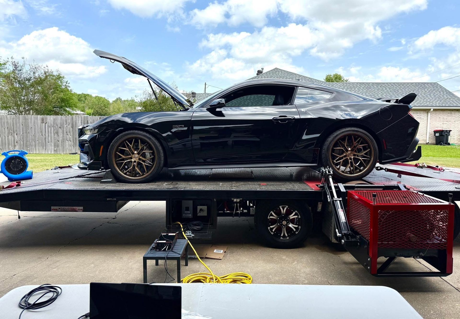 Black sports car on a flatbed trailer with the hood open, under a partly cloudy sky.