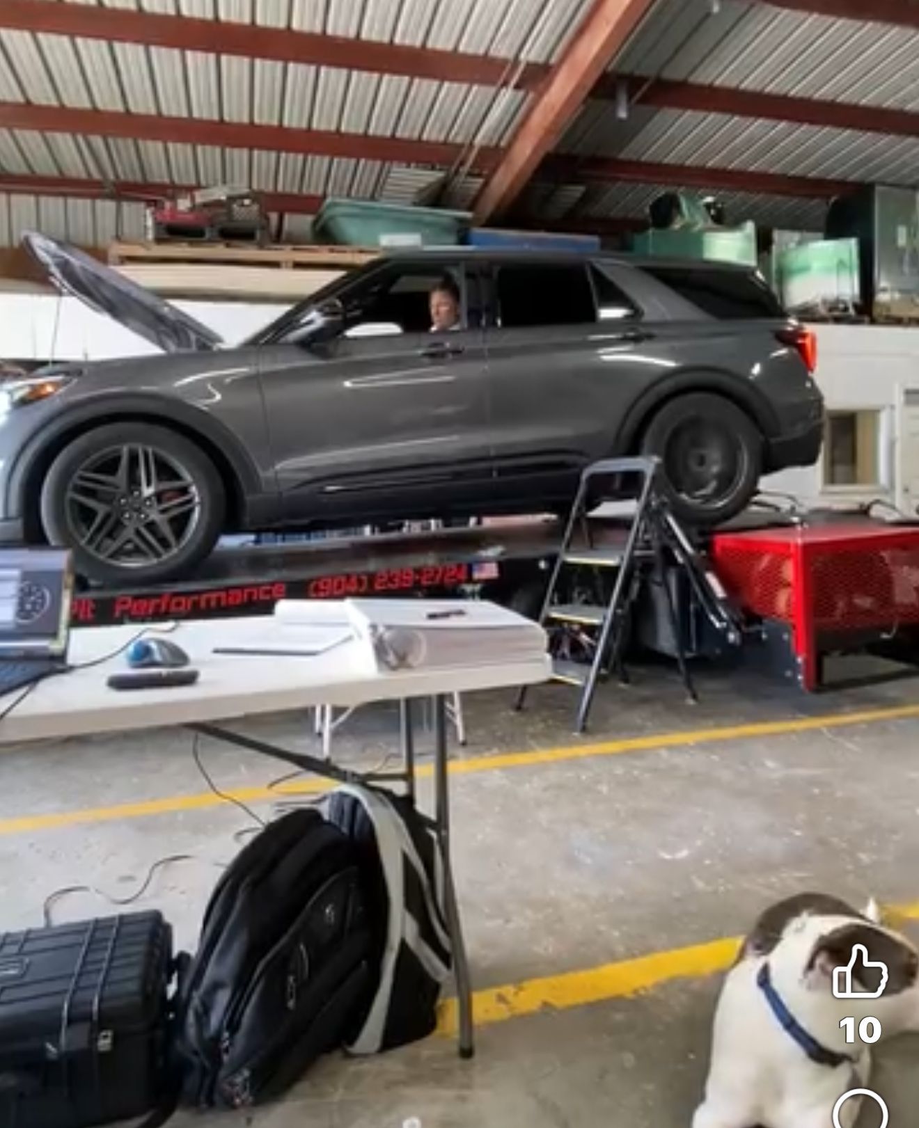 Gray SUV on a lift in a garage with the hood open, someone inside, a dog, and equipment on a table.