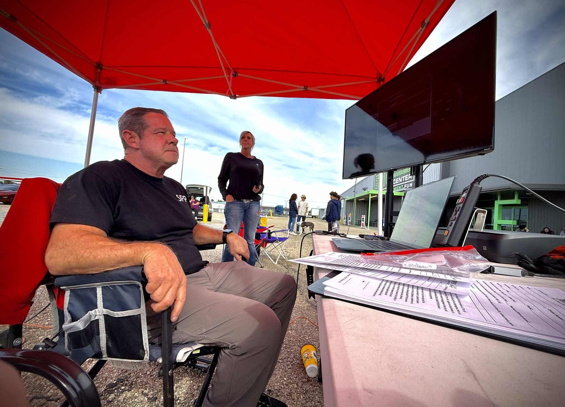 Man seated under red canopy watches screen outdoors. Woman and others in background.
