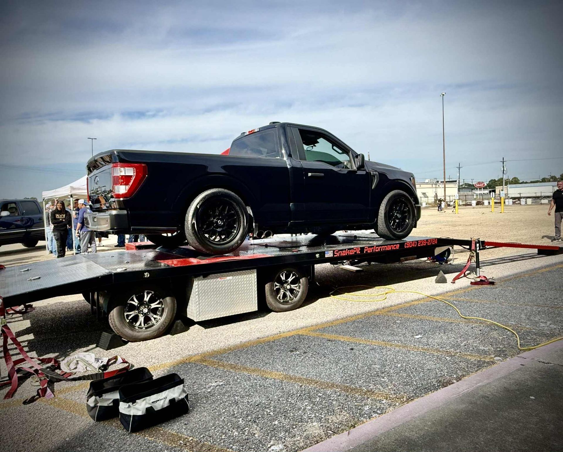 Black pickup truck on a red flatbed trailer, parked on gravel.