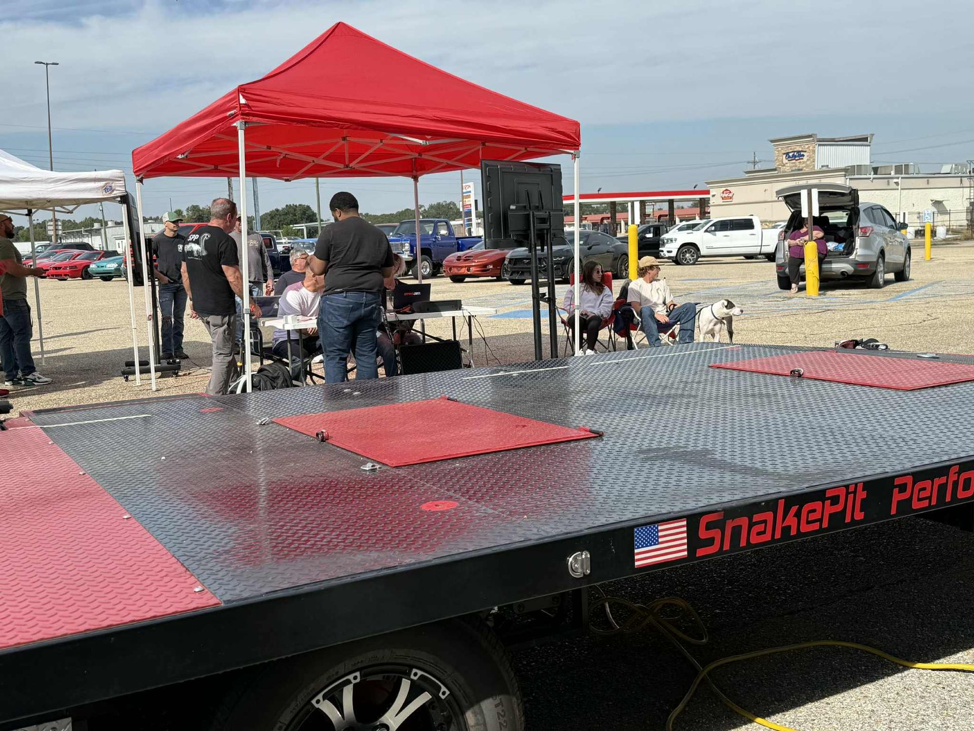 A red-roofed tent set up on a trailer platform, with people gathered around it on a sunny day.