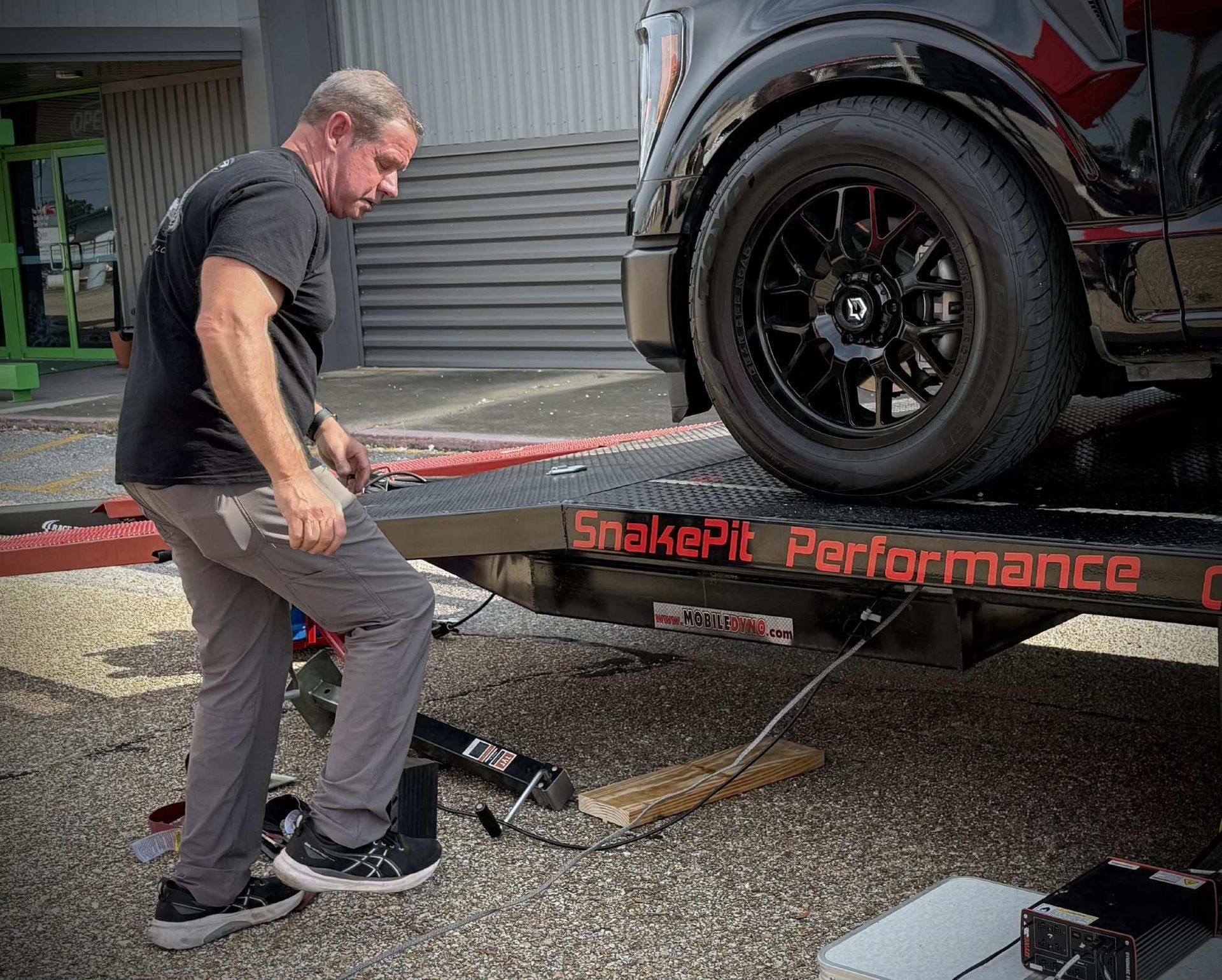 Man lowers a truck from a SnakePit Performance car hauler. Black truck, dark wheels.