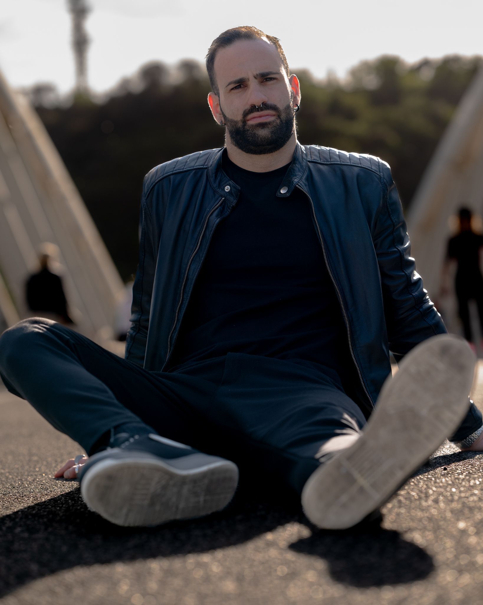 Uomo seduto a gambe incrociate sull'asfalto, con giacca e camicia nere. Sfondo sfocato con ponte.