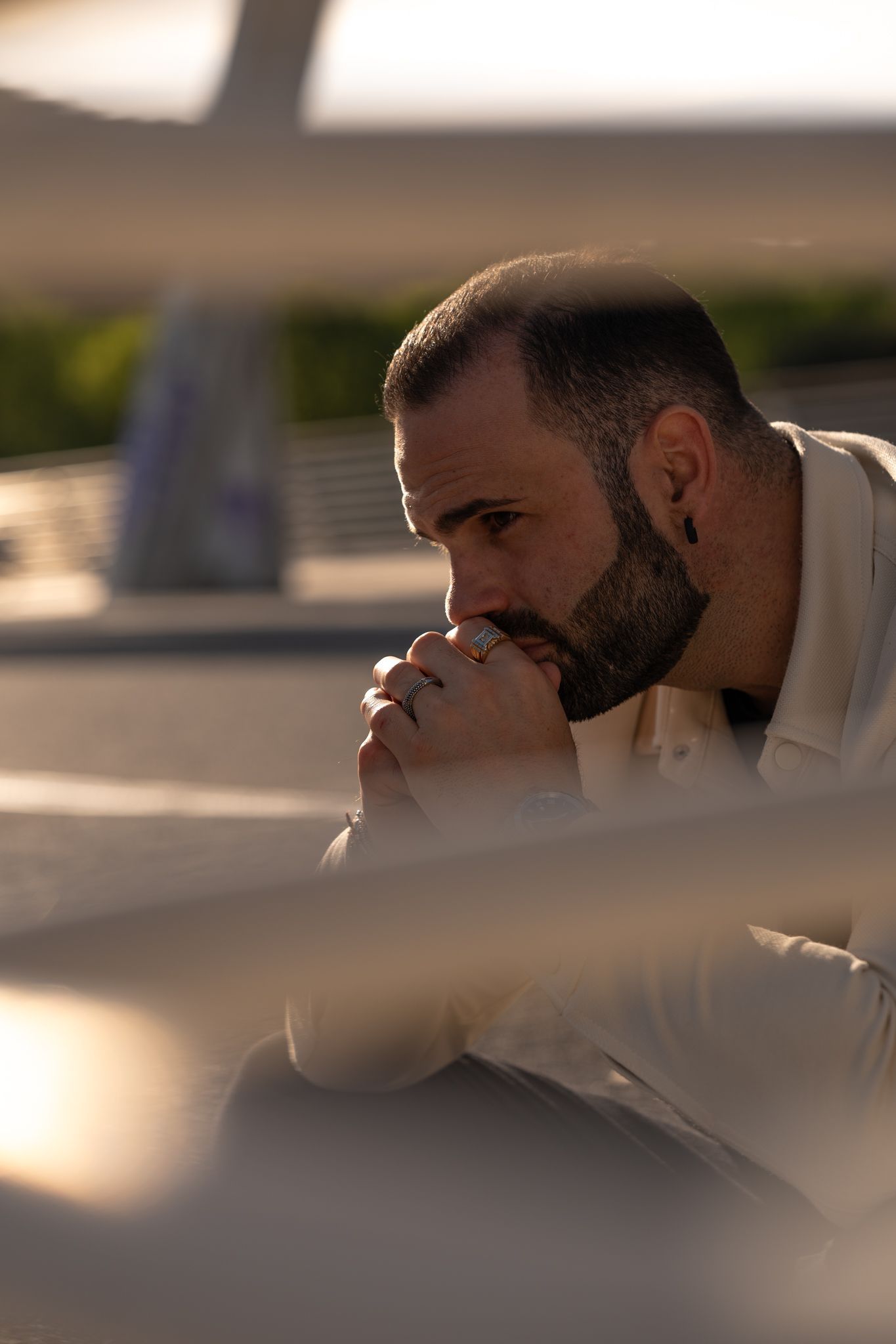 Uomo con la barba, mani giunte, sguardo pensieroso. Seduto all'aperto, forse su un ponte.