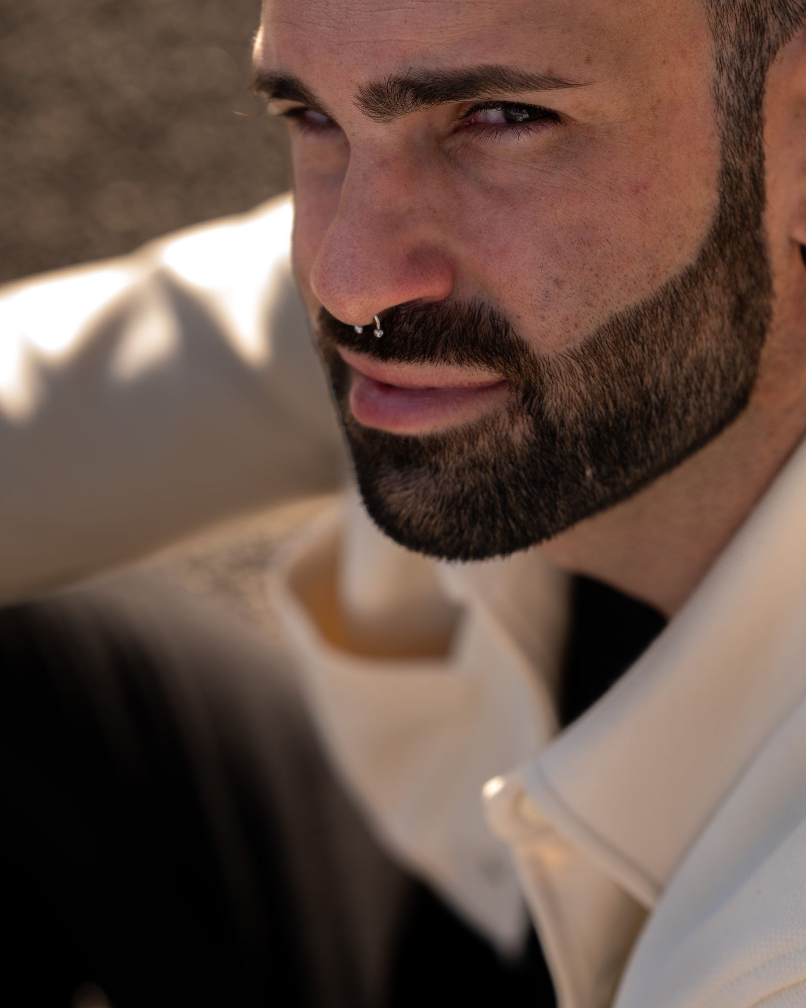 Uomo con barba e anello al naso, che indossa una giacca color crema, guarda fuori dall'inquadratura.