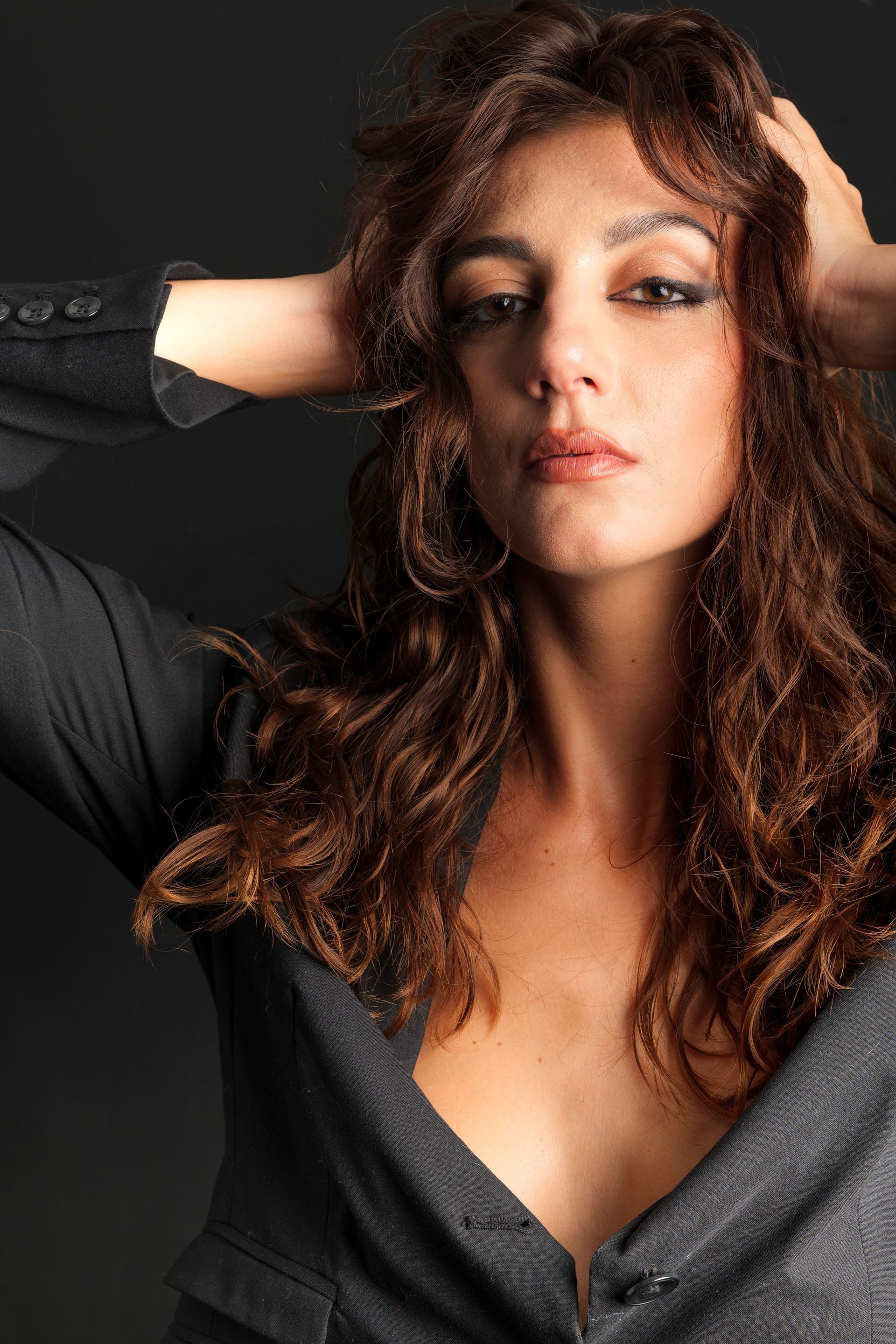 Donna con capelli castani ondulati, mani sulla testa, giacca nera, espressione neutra.
