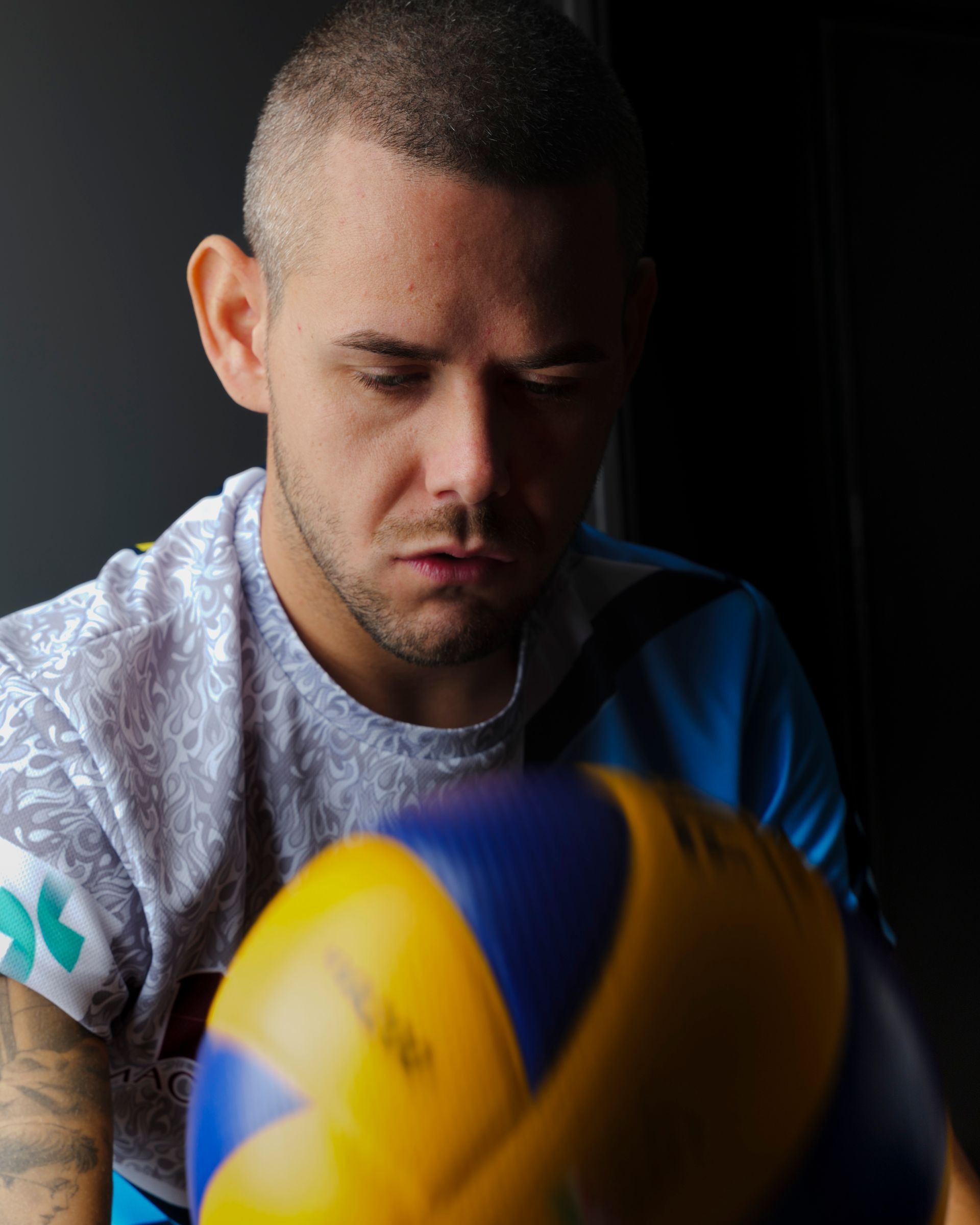 Uomo con la testa rasata che tiene in mano un pallone da pallavolo, guardando pensieroso verso il basso. In un interno, davanti a uno sfondo scuro.