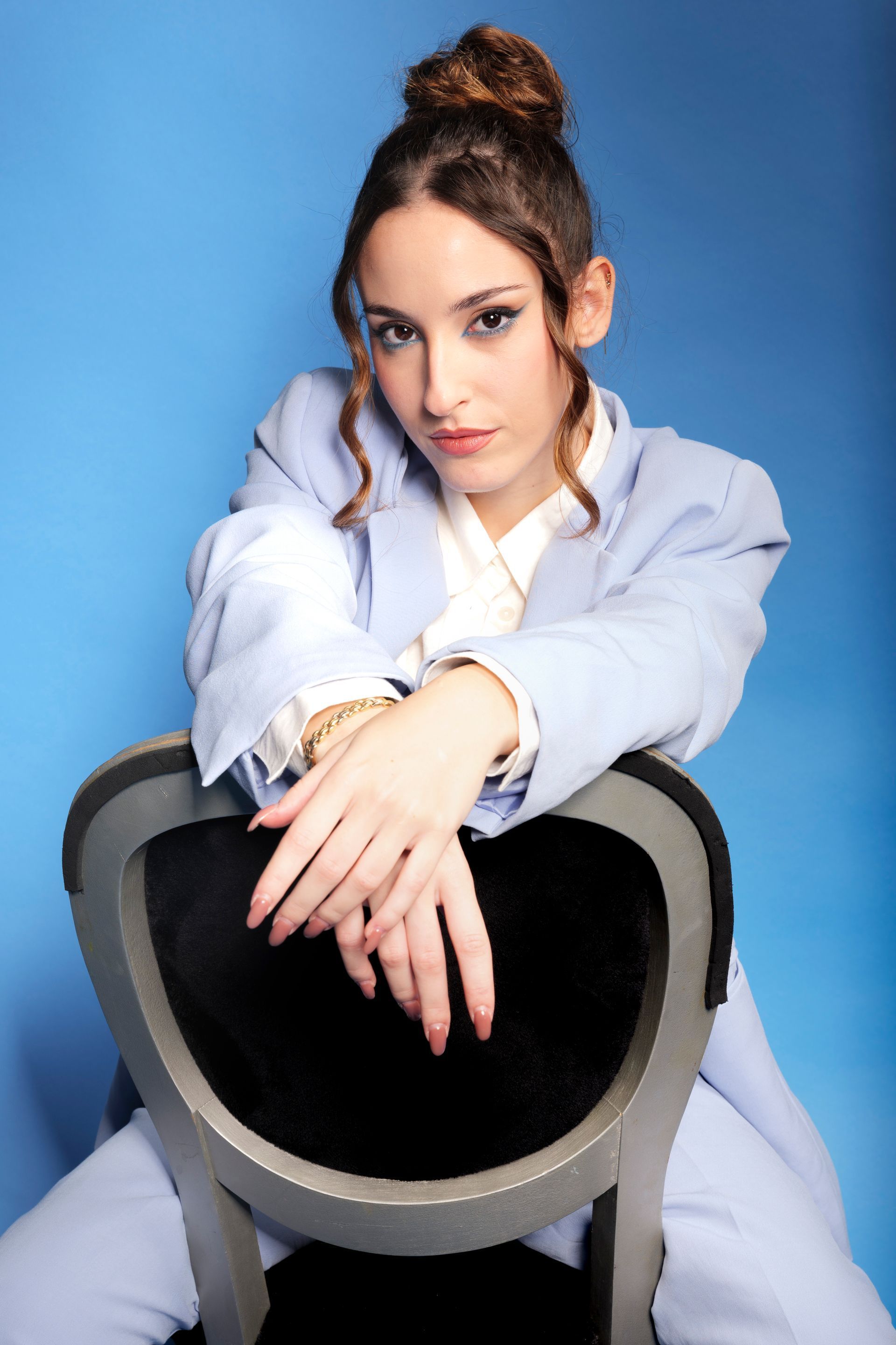 Una donna in abito azzurro, con i capelli raccolti in uno chignon, è appoggiata a una sedia su uno sfondo blu.