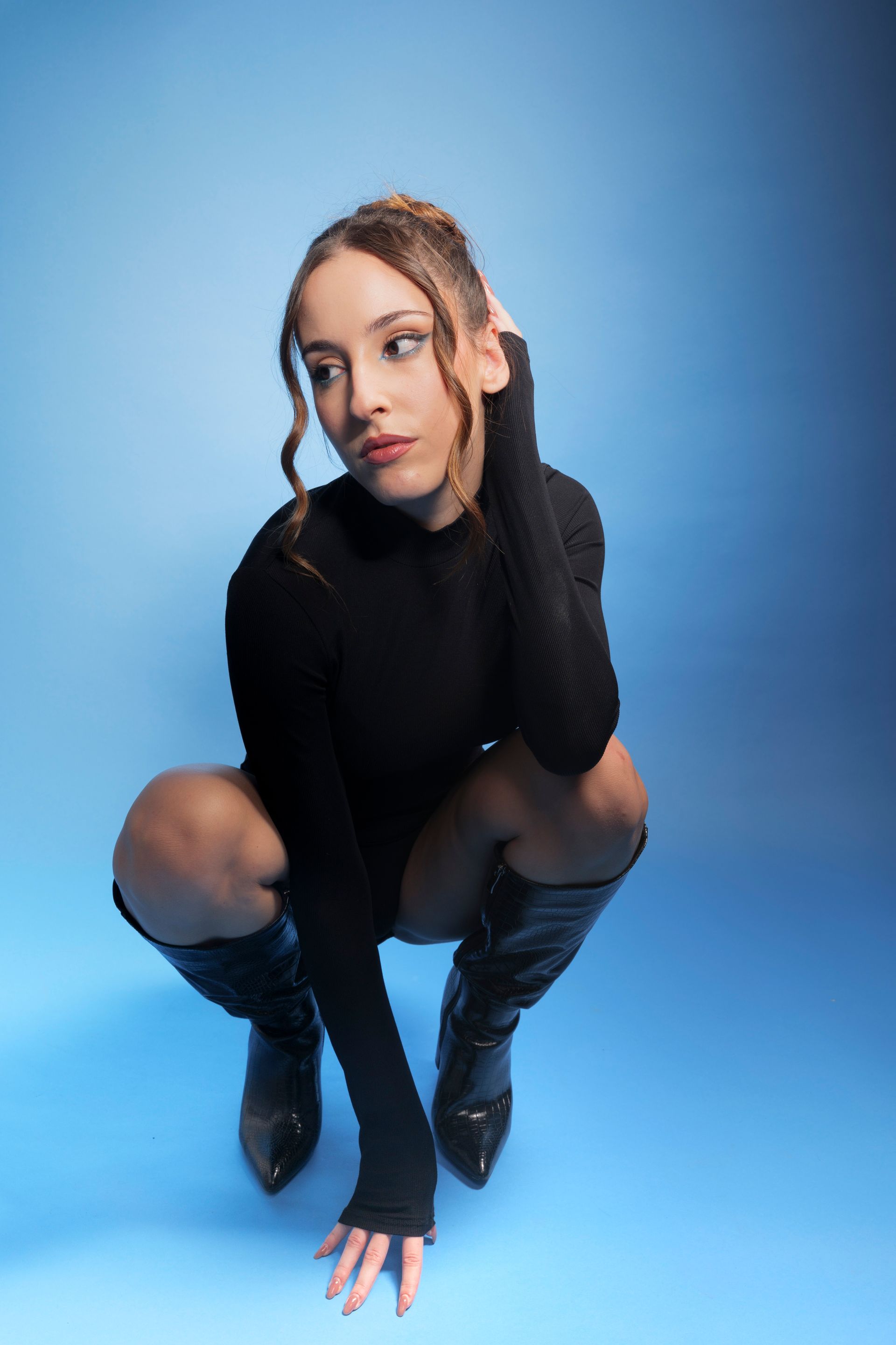 Una donna in abito nero e stivali è accovacciata su sfondo blu e guarda altrove.