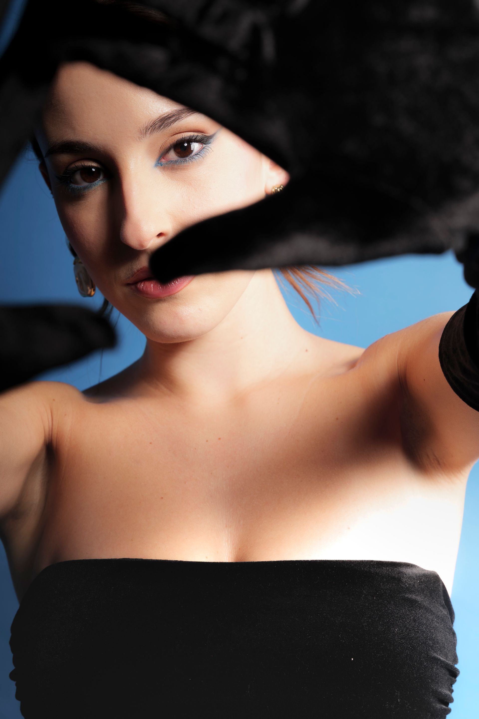 Donna incorniciata da mani guantate, che indossa un top nero senza spalline, su sfondo blu.