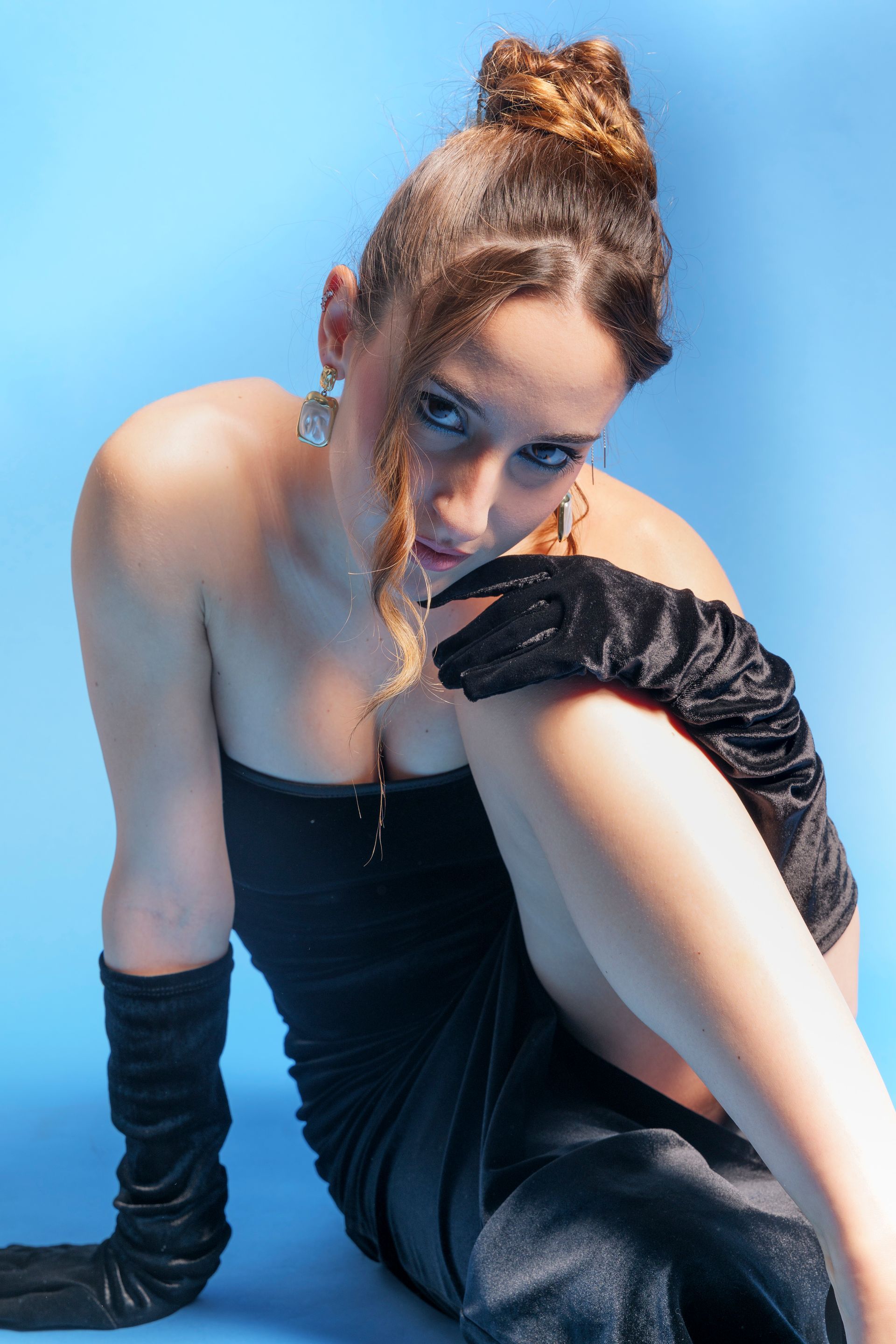 Una donna in abito da sera nero posa su uno sfondo blu, indossando guanti e orecchini.