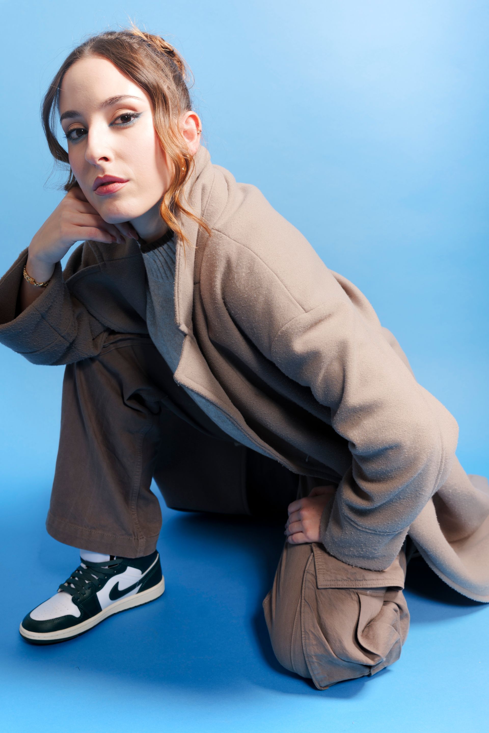 Un giovane in abito marrone si accovaccia e guarda in camera. Indossa scarpe da ginnastica, cappotto e si trova su uno sfondo blu.