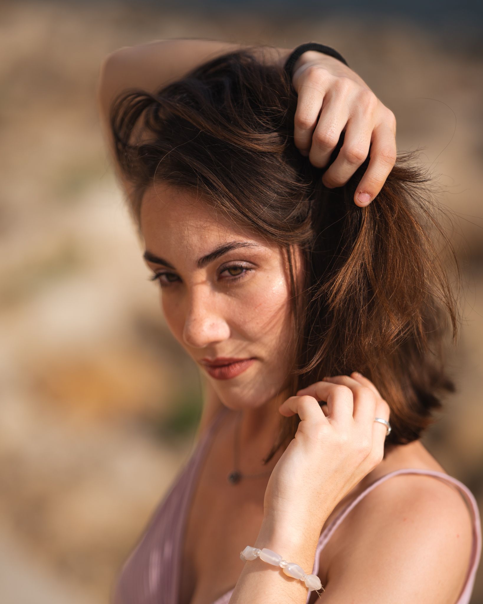 Una donna con i capelli castani e una maglietta rosa guarda verso il basso, con le mani tra i capelli, all'aperto, alla luce del giorno.