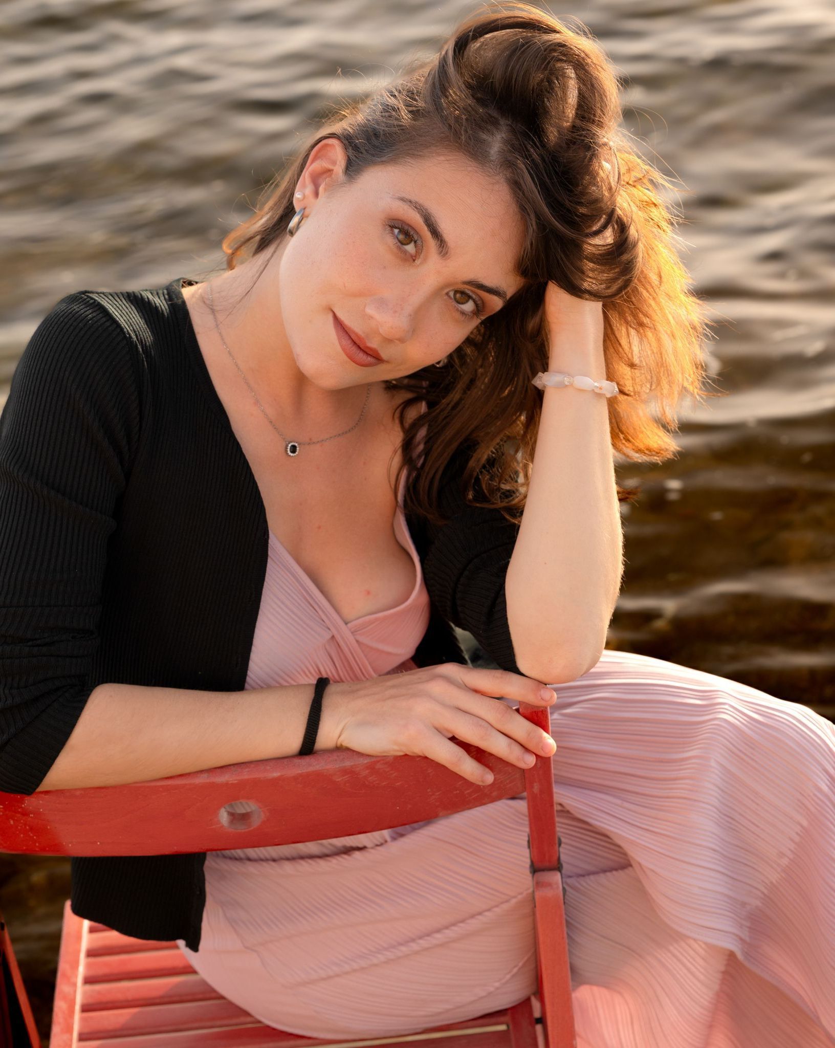 Donna con abito rosa e maglione nero, appoggiata a una sedia rossa in riva all'acqua.