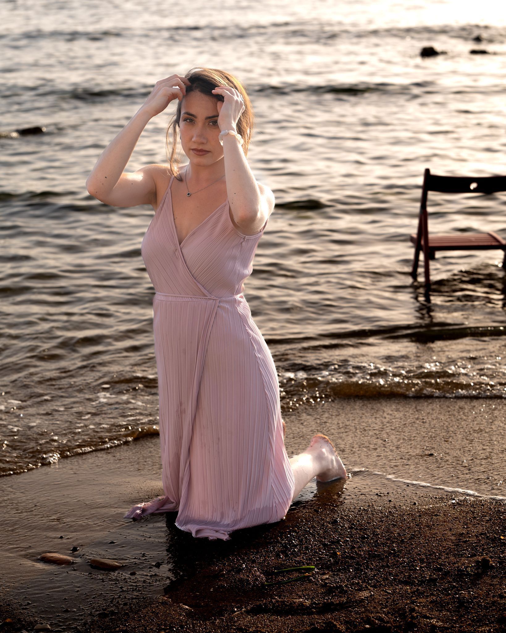 Una donna si inginocchia sulla spiaggia con un abito rosa, le mani nei capelli, una sedia lì vicino, acqua, tramonto.