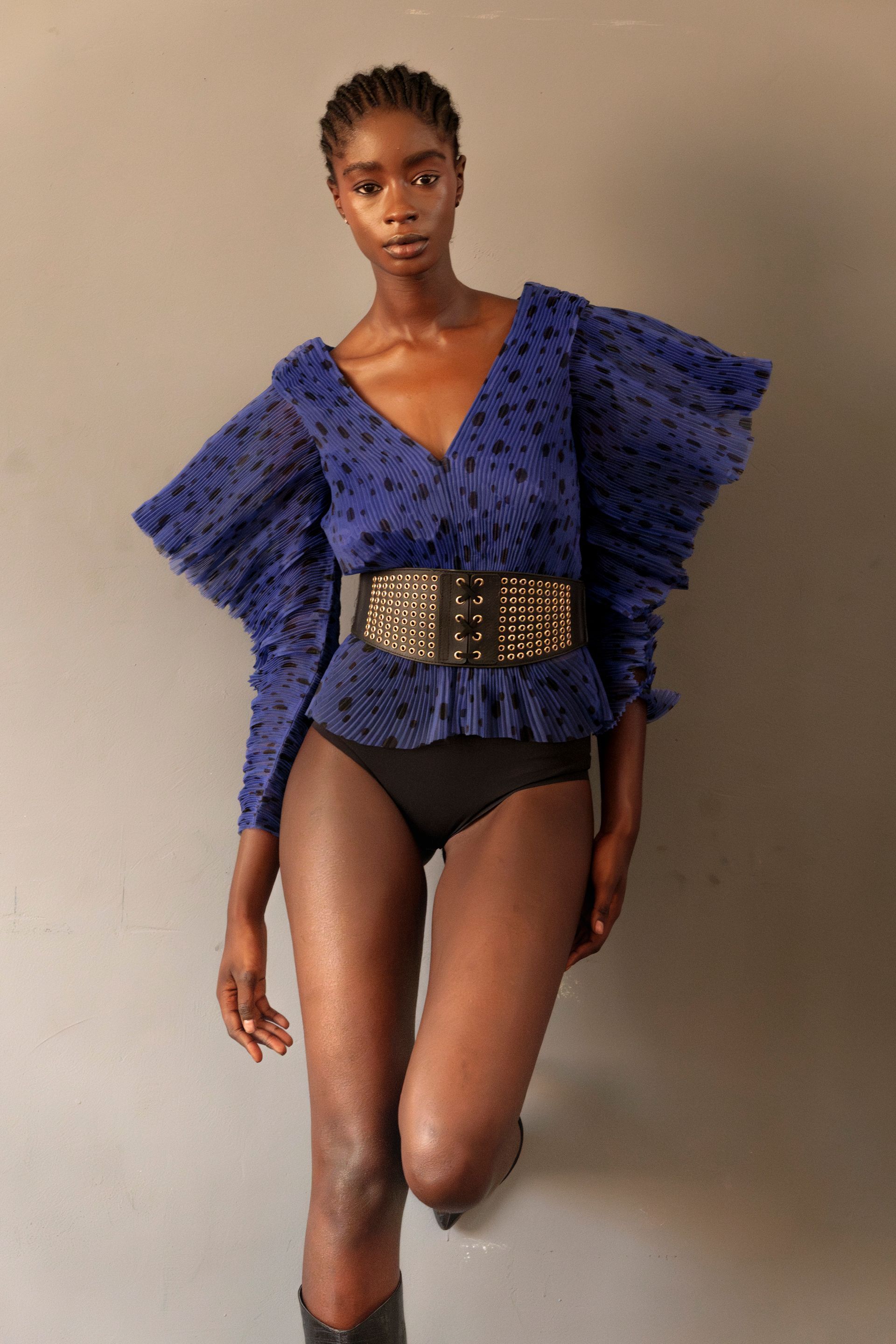 Donna con top blu con maniche a sbuffo, pantaloni neri e cintura larga, appoggiata a un muro grigio.